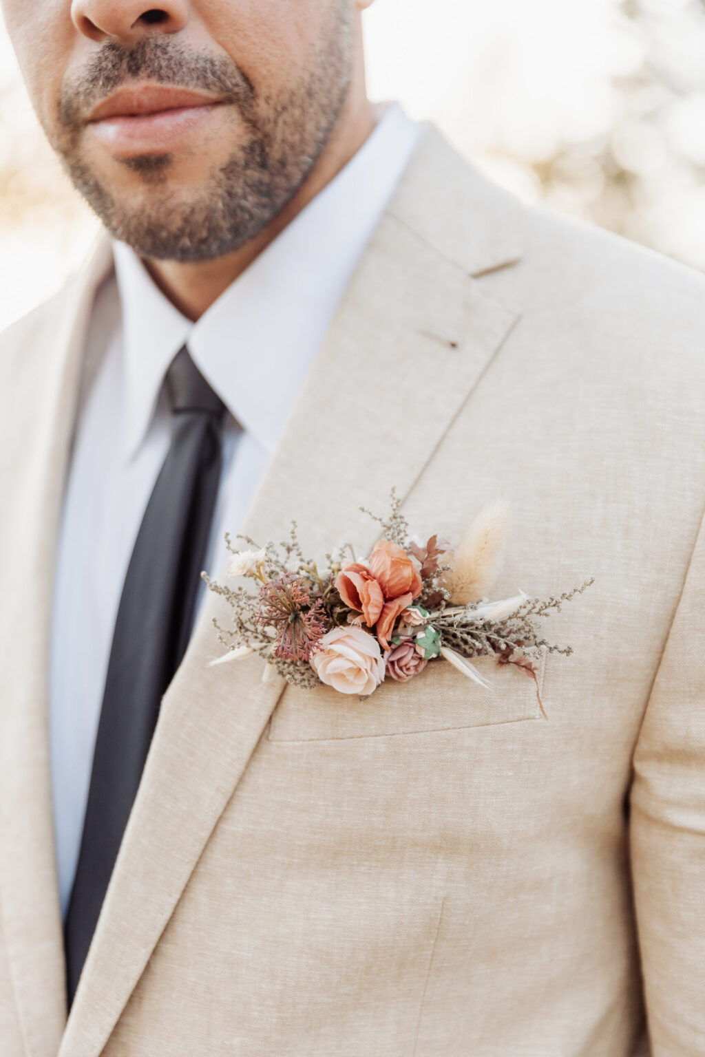 Elopement Wedding A man is shown from the shoulders up wearing a light beige suit jacket, white shirt, and black tie. He has a dark beard and mustache. A boutonniere made of pink, cream, and orange flowers with greenery is pinned to his left lapel. The softly blurred background hints at an intimate elopement setting. Elopements Inc