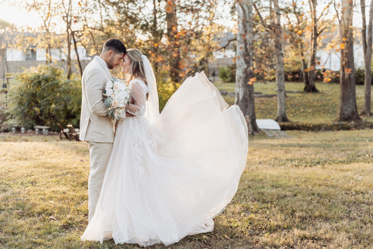 Elopement Wedding A bride in a flowing white gown and veil embraces a groom in a beige suit in a grassy, wooded area. The groom holds her waist, and the bride's dress fans out as they gaze at each other tenderly. Sunlight filters through the trees, creating a warm, romantic ambiance perfect for their intimate elopement. Elopements Inc