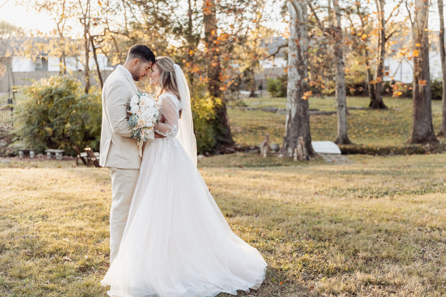 Elopement Wedding A bride and groom stand closely together in an outdoor, wooded area. The groom wears a light-colored suit, while the bride is in a long, flowing white gown with a veil, holding a bouquet of flowers. They lean their foreheads together, savoring the intimacy of their elopement amidst trees with autumn leaves. Elopements Inc