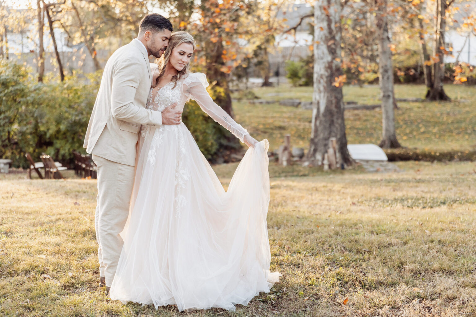 Elopement Wedding A couple stands in a sunlit autumnal garden, posing for a photo. The woman wears a long, flowing white wedding dress with lace details and long sleeves, holding her dress with one hand. The man is dressed in a light beige suit, gently holding the woman around the waist. Trees with fall foliage are in the background—a perfect scene for elopements. Elopements Inc