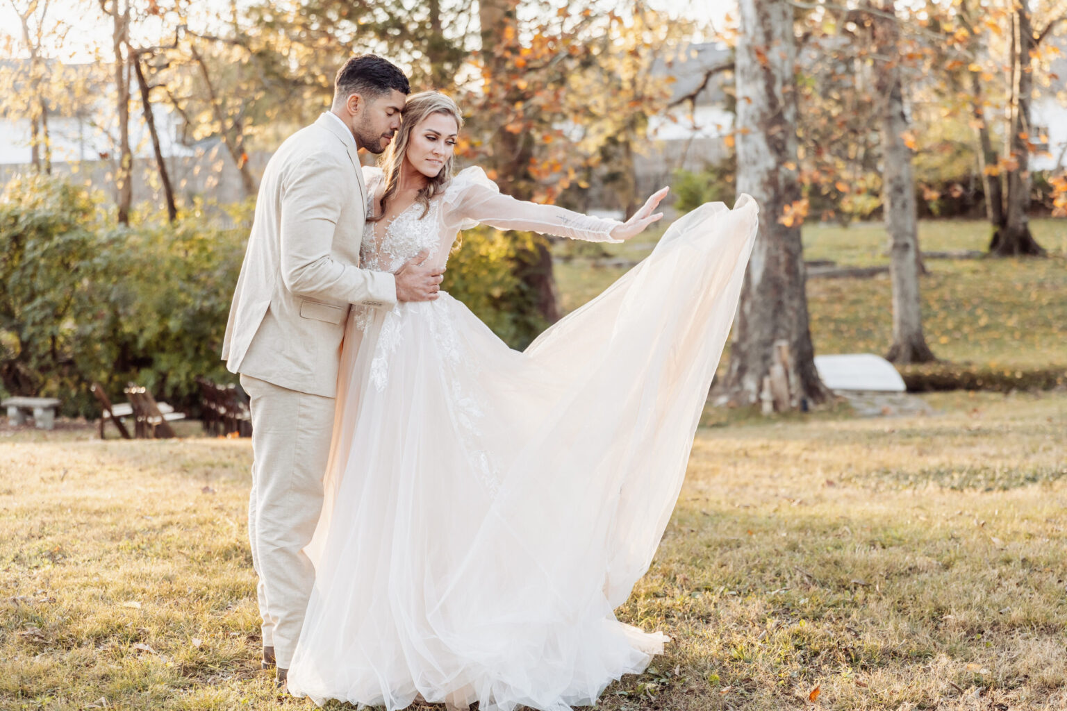 Elopement Wedding A bride and groom stand on a grassy lawn surrounded by trees, ready to elope. The groom, in a beige suit, holds the bride around her waist. The bride, in a long, flowing white gown with lace details, extends her arm out, lifting one side of her dress. The background features trees with fall foliage. Elopements Inc