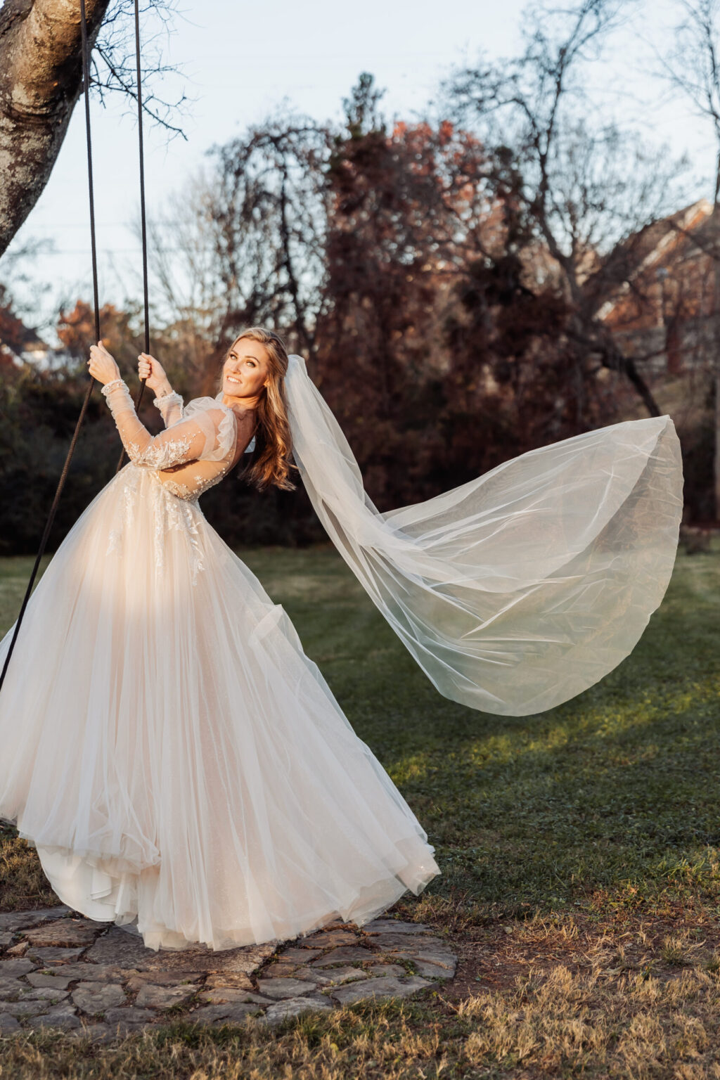 Elopement Wedding A joyful bride in a long-sleeved, lace and tulle wedding dress swings beneath a tree in a sunlit garden. Her long veil flows elegantly in the air, capturing the movement. The grassy area and autumnal trees in the background add a picturesque, romantic feel to this intimate elopement scene. Elopements Inc