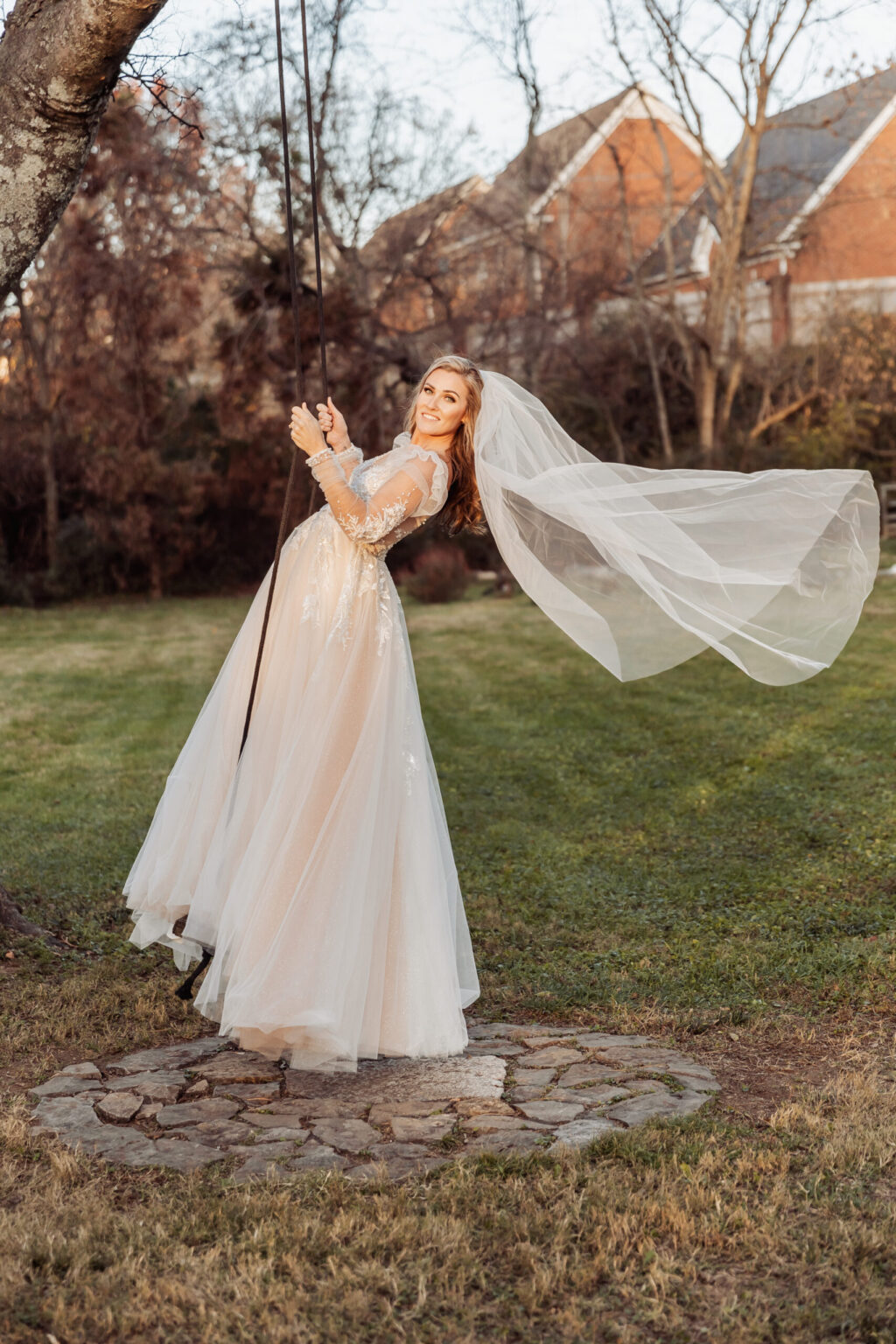 Elopement Wedding A bride in a long, flowing white wedding dress with lace sleeves and a veil is joyfully swinging on a tree swing. She is outdoors on a grass lawn with some brick houses and leafless trees visible in the background. The sunlight enhances the serene and joyful atmosphere of her intimate elopement. Elopements Inc