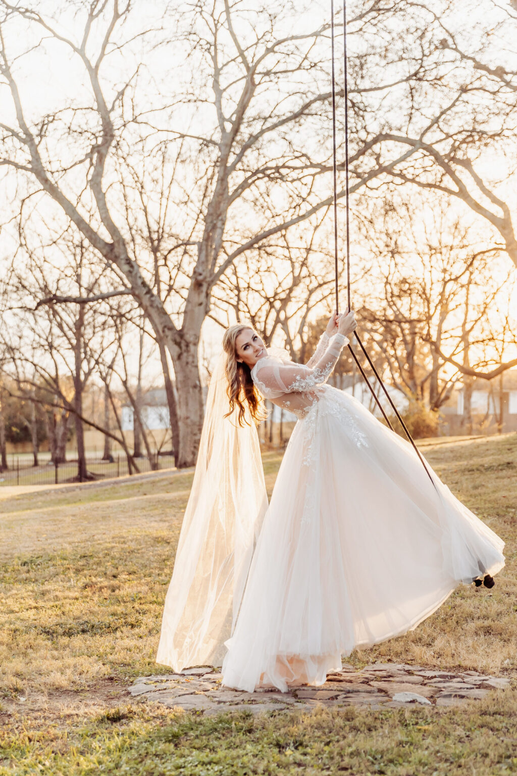 Elopement Wedding A bride in a flowing white gown and long veil is joyfully swinging on a rope swing at sunset, celebrating her elopement. The scene is set in a serene, open area with a backdrop of bare trees and a softly glowing sky. Her gown has lace details and a sheer overlay, adding elegance to the moment. Elopements Inc