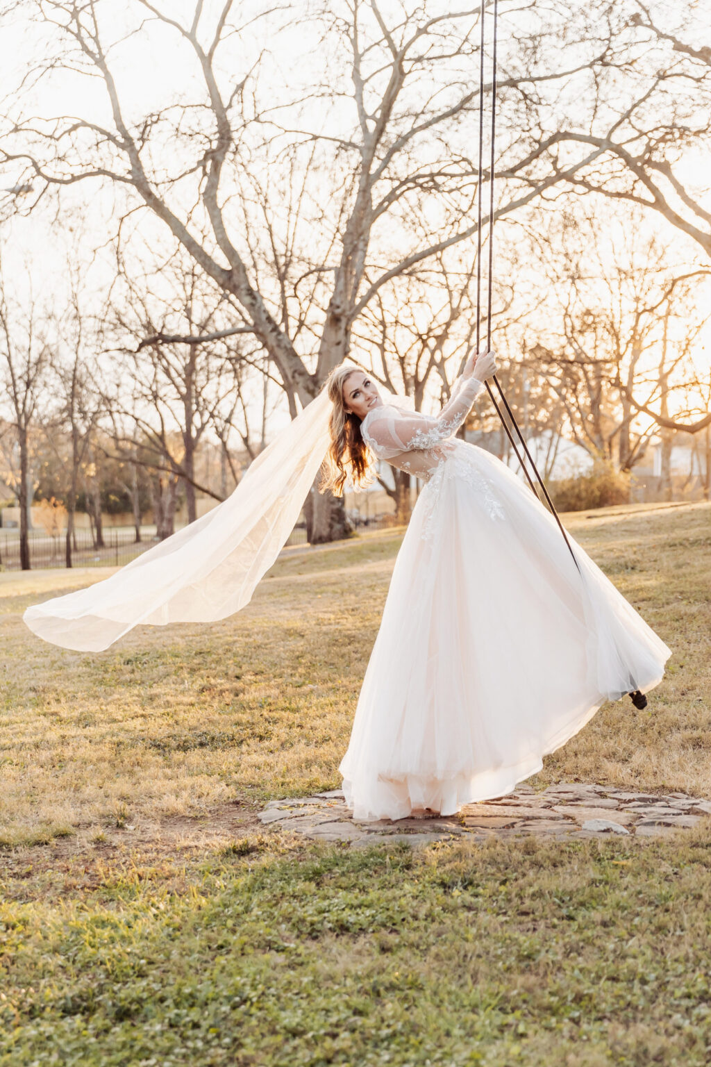 Elopement Wedding A woman in a white wedding dress, with a long, flowing veil, stands on a swing in an outdoor setting. She is leaning back with one arm extended and holding onto the swing ropes. The leafless trees and sunset create a warm glow, perfect for an intimate elopement scene. Elopements Inc