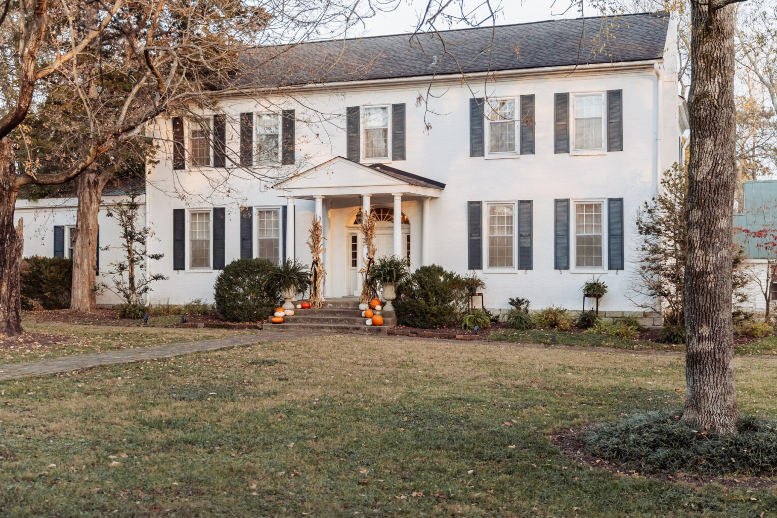 Elopement Wedding A two-story white colonial house features black shutters and a neatly manicured front lawn. The entrance is adorned with a central portico supported by columns, with a small porch decorated with pumpkins. Leafless trees and seasonal greenery hint at late autumn, making it a picturesque backdrop for elopements. Elopements Inc