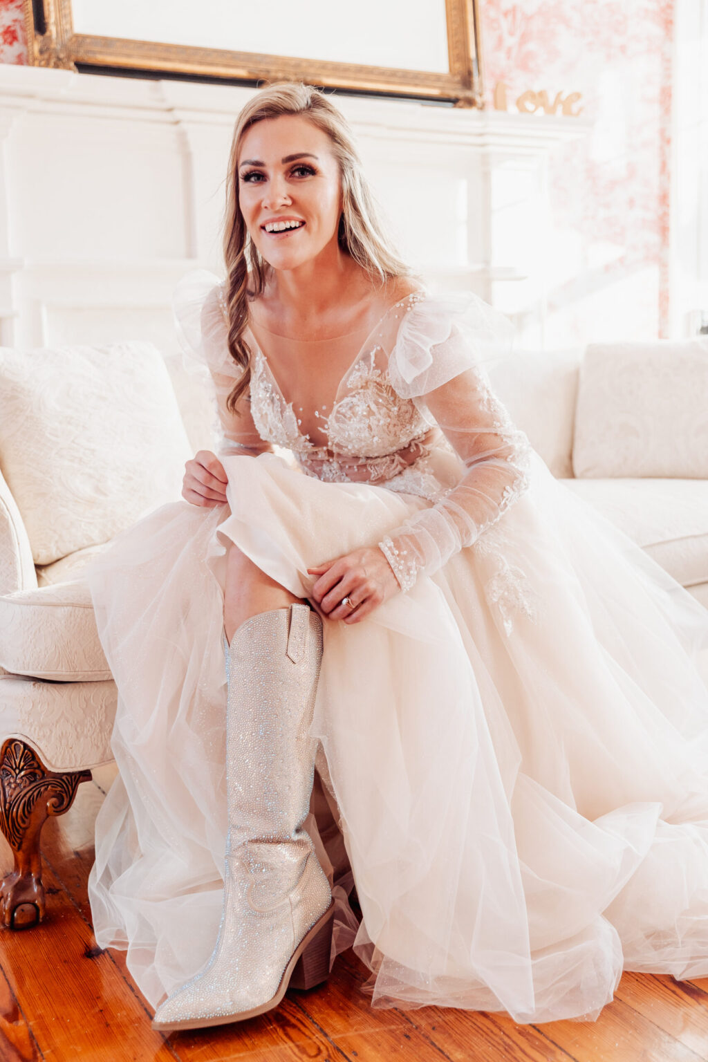 Elopement Wedding A smiling bride with long blonde hair sits on a white sofa, wearing a white wedding dress with sheer lace sleeves and a plunging neckline. She is putting on sparkly silver cowboy boots, perhaps ready for an adventurous elopement. The background features a white wooden mantle adorned with the word "LOVE. Elopements Inc