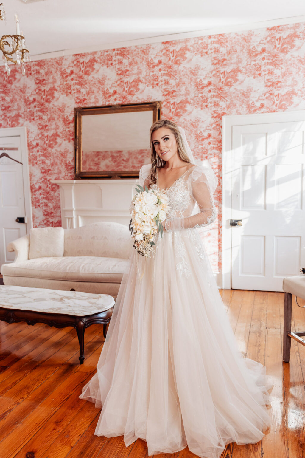 Elopement Wedding A bride in a long, flowing white wedding dress with lace sleeves and a sheer overlay stands in a room with red-patterned wallpaper, ready for an intimate elopement. She holds a bouquet of white flowers. Behind her, there is a white couch, a marble coffee table, and a large mirror above a white fireplace. Sunlight illuminates the room. Elopements Inc