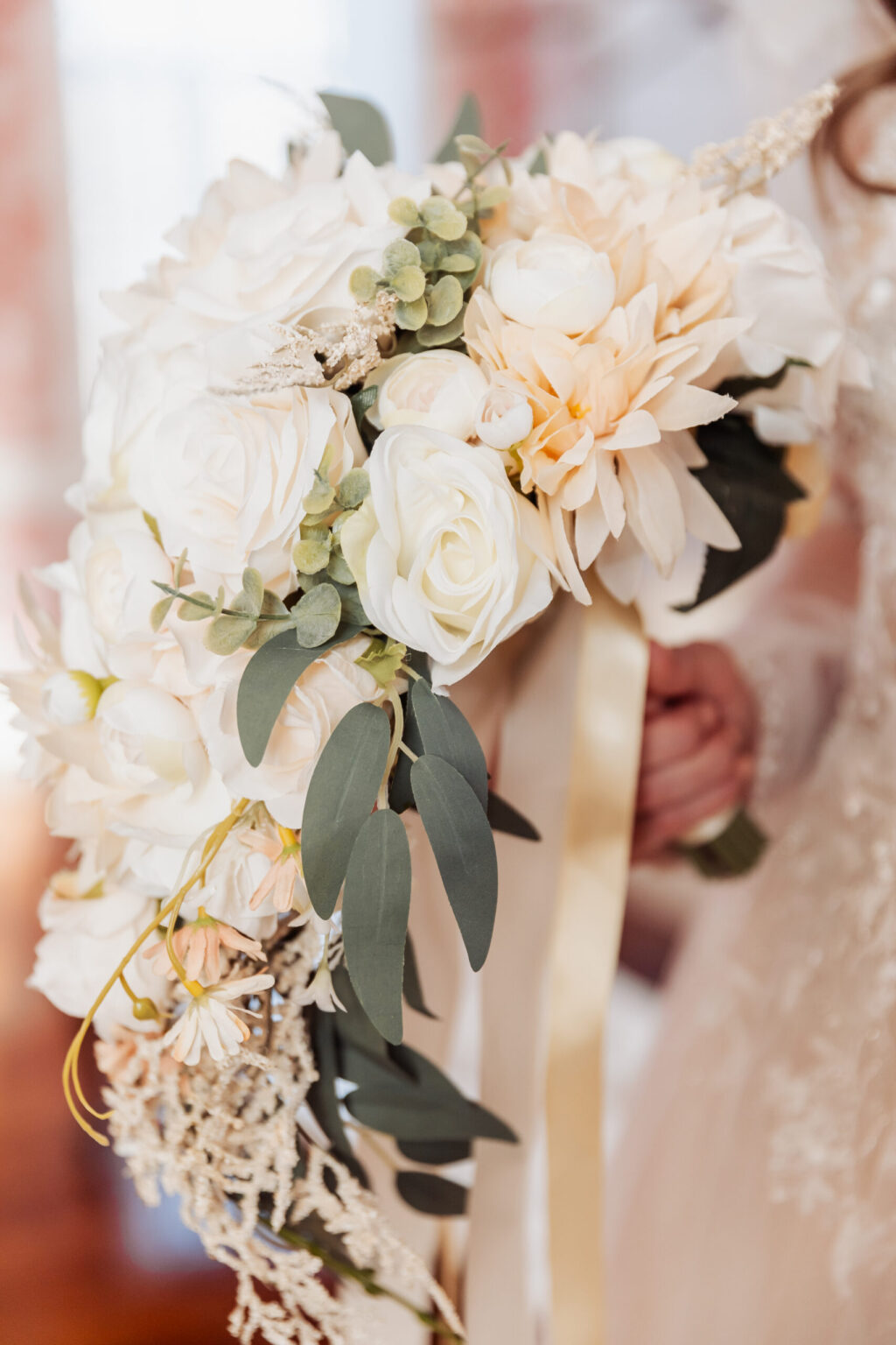 Elopement Wedding A close-up photograph of a bride holding a cascading bouquet, perfect for an intimate elopement. The bouquet features a mix of white and pale peach flowers, including roses and dahlias. Greenery interspersed among the flowers and a ribbon trailing down enhance the elegant arrangement. The bride is dressed in a lace gown. Elopements Inc