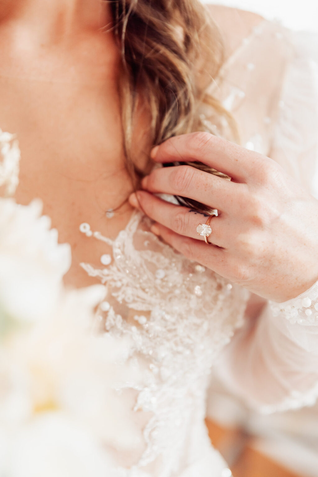 Elopement Wedding A close-up image of a bride's hand touching her chest. She is wearing a delicate, lacy wedding dress. On her ring finger is a diamond ring. Her hair cascades in loose waves over her shoulder. The focus is on her hand, the ring, and the intricate details of her dress, perfect for an elopement. Elopements Inc