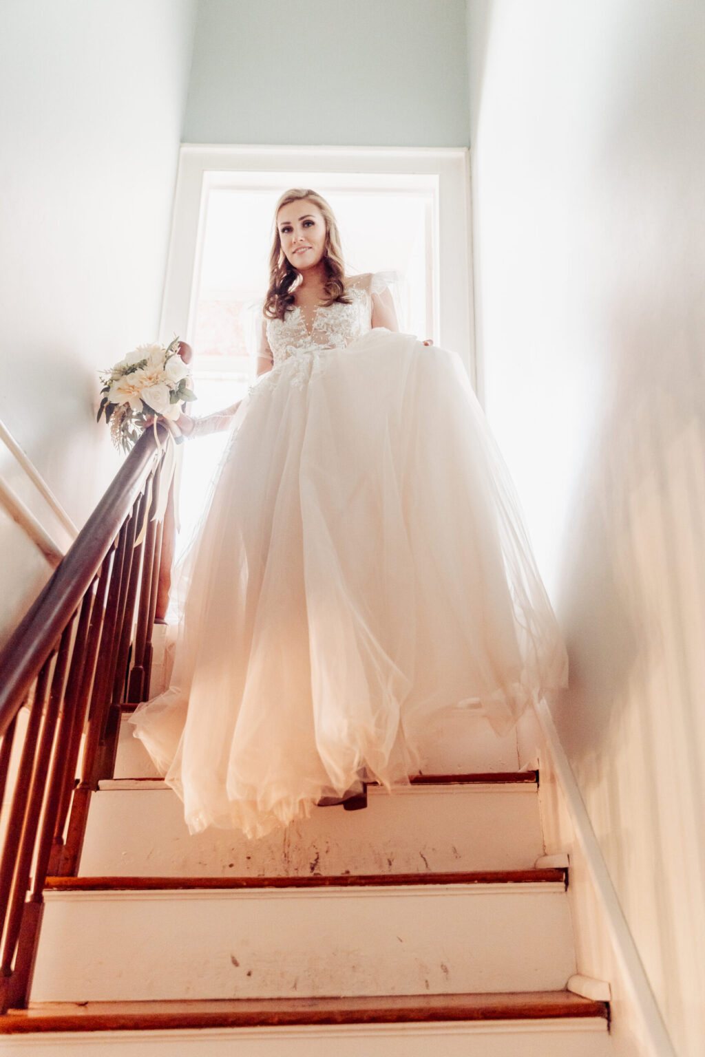 Elopement Wedding A bride in a flowing white wedding dress descends a wooden staircase, holding a bouquet of white flowers. She is captured in natural light from a large window behind her, creating a soft, romantic ambiance perfect for an elopement. The staircase is narrow, with white steps and wooden railings. Elopements Inc