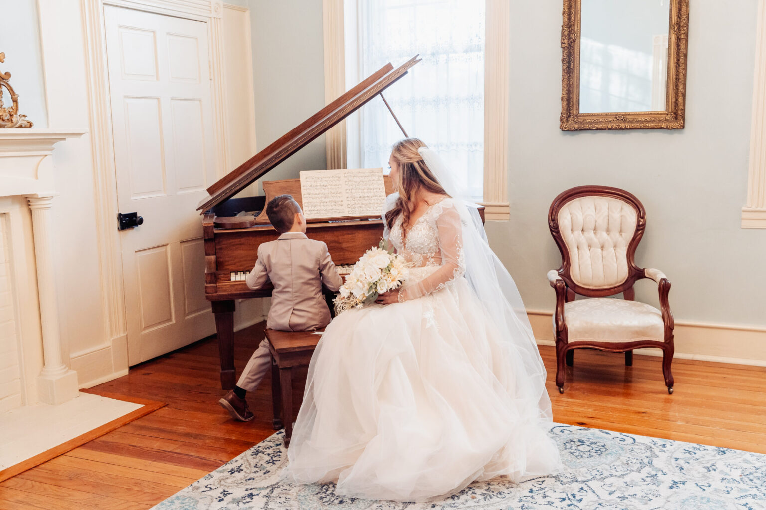 Elopement Wedding A bride in a white wedding dress and veil, just after an intimate elopement, sits beside a young boy playing a grand piano. Holding a bouquet of white flowers, she gazes toward the sheet music. The room features a cream-colored wall, a decorative fireplace, a large window, and elegant furniture. Elopements Inc