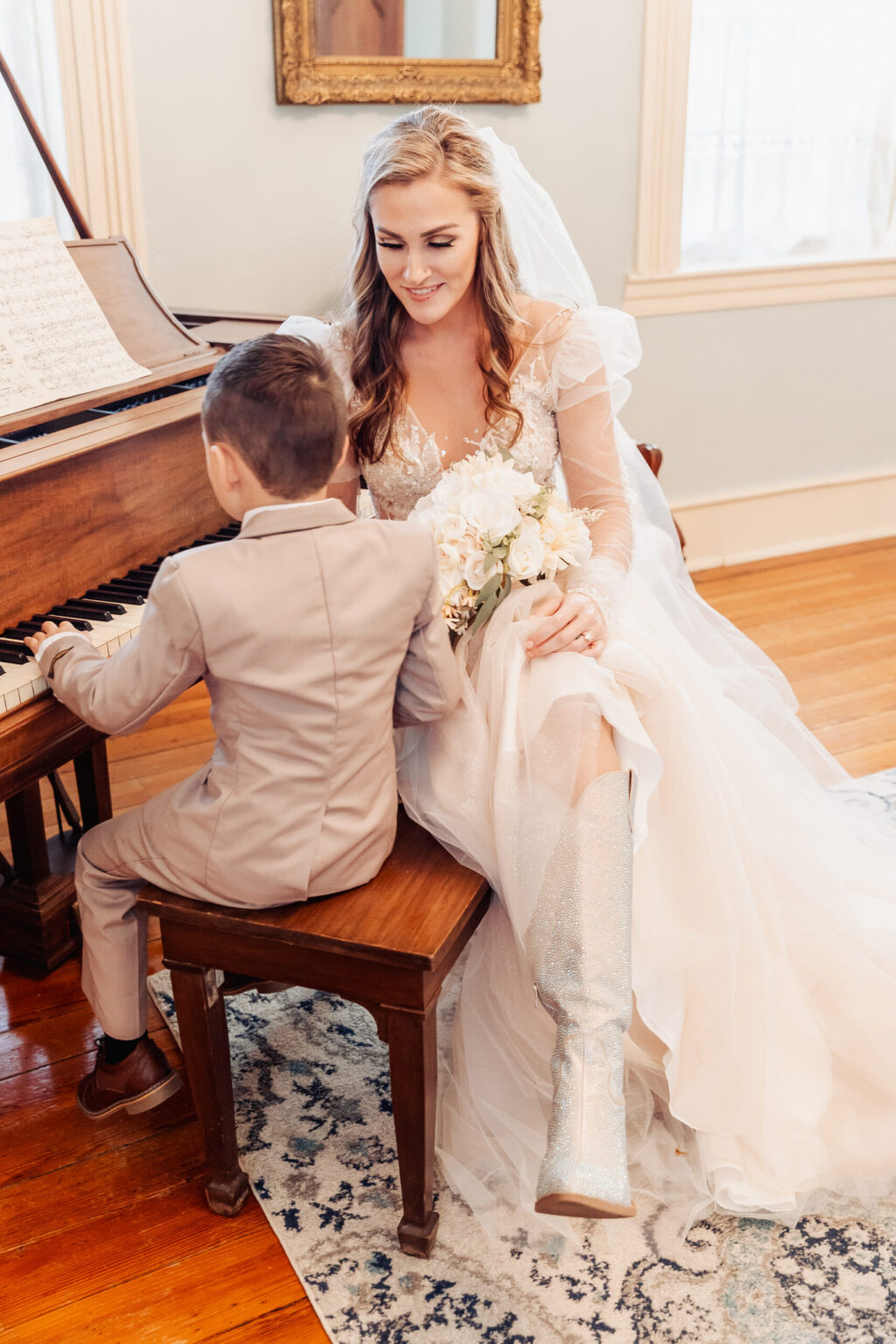 Elopement Wedding A bride in a white wedding dress and sparkly boots sits beside a young boy playing the piano, capturing a moment filled with joy during their intimate elopement. She holds a small bouquet of white flowers, smiling down at the boy. They are in a warmly lit room with wooden floors, a patterned rug, and sheer curtains by the window. Elopements Inc