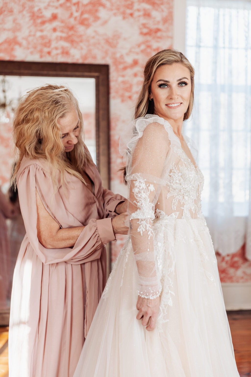 Elopement Wedding A bride in a white, long-sleeved, lace wedding dress stands smiling as an older woman in a blush pink dress adjusts the back of her gown. The background has pink floral wallpaper, a mirror, and a window with sheer curtains allowing natural light to filter into the room—perfect for intimate elopements. Elopements Inc