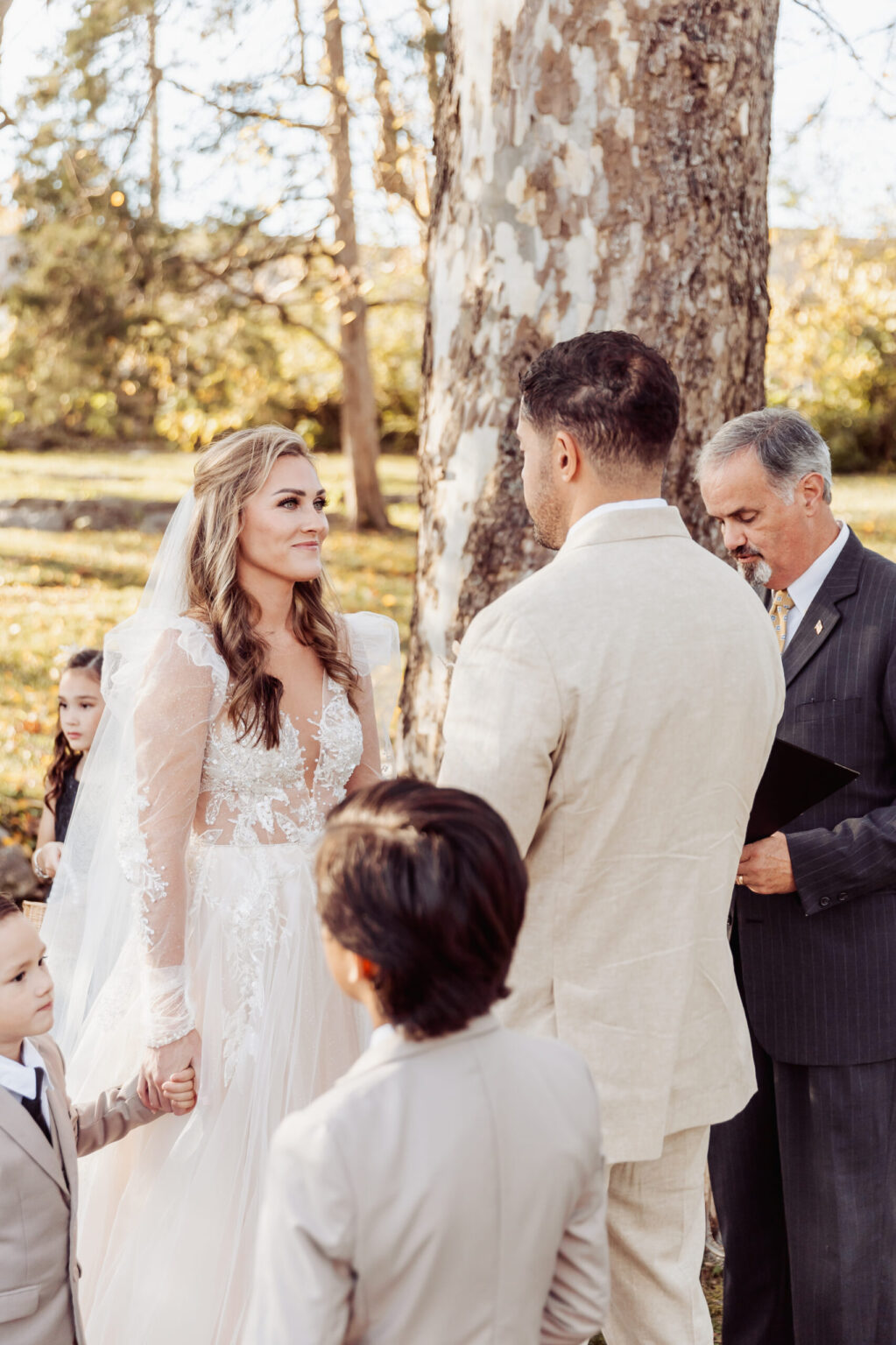 Elopement Wedding A bride and groom stand facing each other, holding hands during an intimate outdoor elopement. The bride wears a white lace gown and veil, while the groom wears a beige suit. An officiant in a gray suit stands nearby holding a black book. Two children dressed in formal attire are visible in the foreground. Elopements Inc