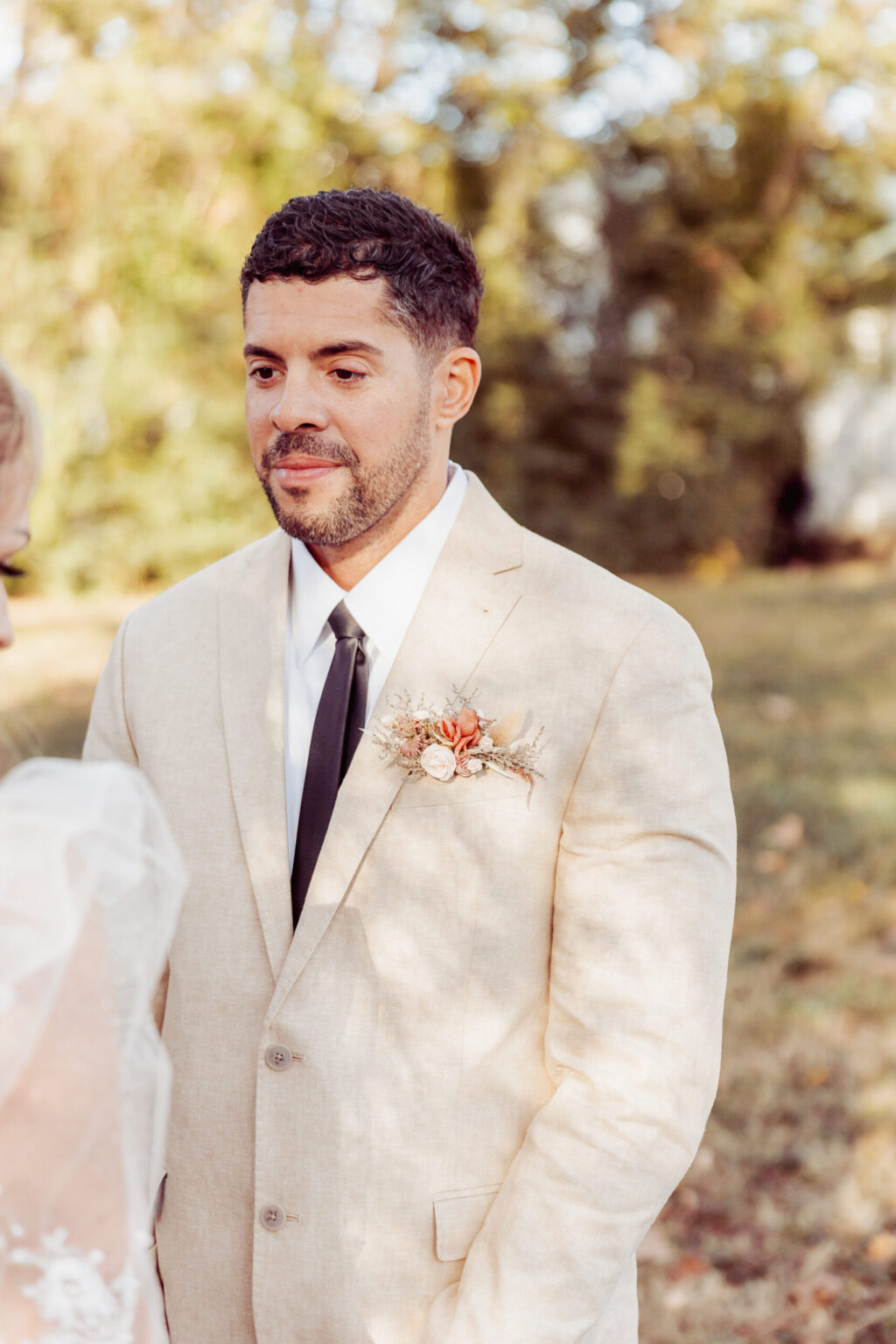 Elopement Wedding A man wearing a beige suit with a white shirt and black tie stands outdoors with greenery in the background. He has short dark hair and a neatly trimmed beard. His suit jacket features a boutonniere with orange and beige flowers. A part of another person, likely the bride, hints at an intimate elopement. Elopements Inc