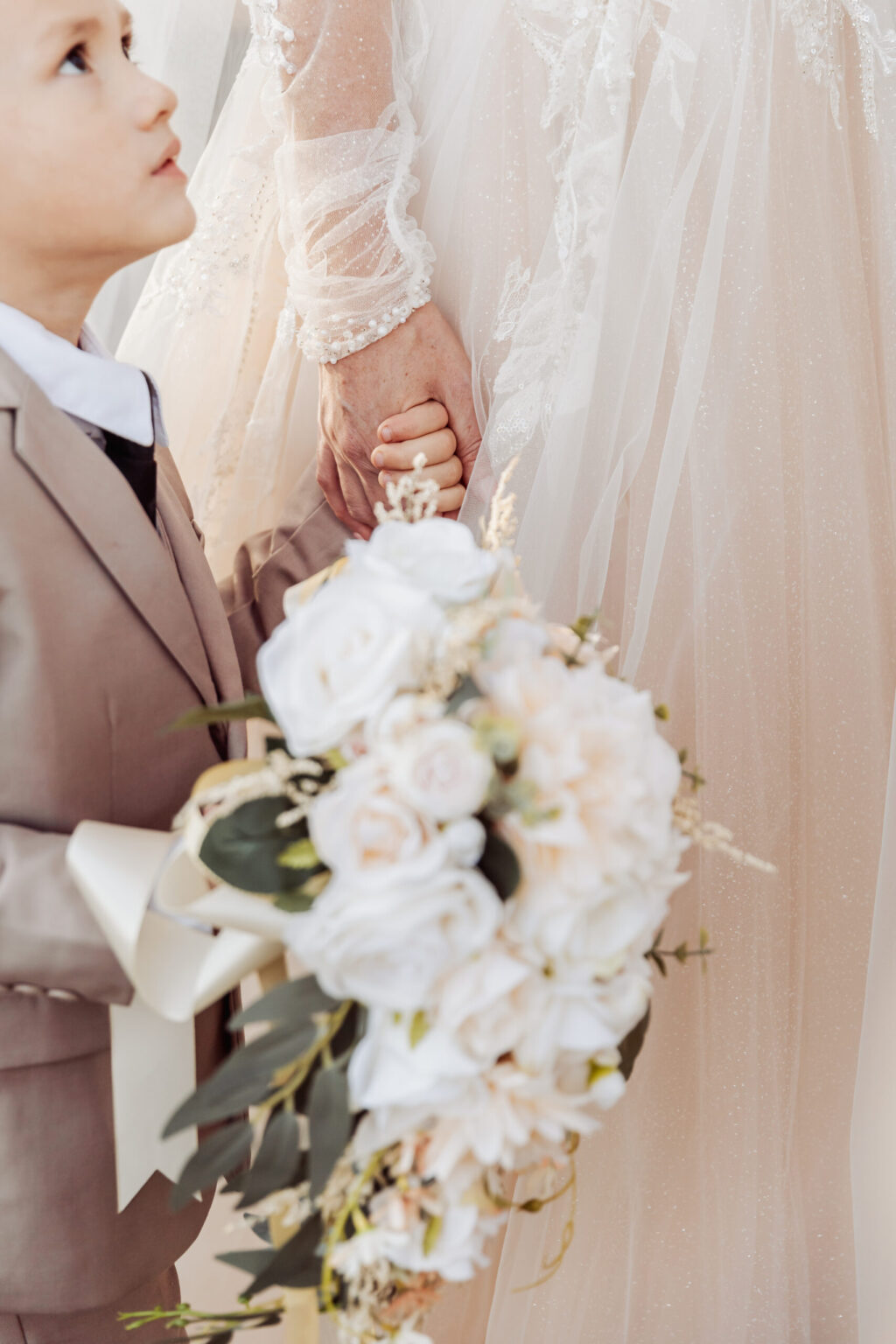 Elopement Wedding A young boy in a light brown suit and white shirt holds a bouquet of white flowers. He is looking up while holding the hand of an adult woman in a lace wedding dress, whose face is not visible. The setting appears to be a formal occasion, perhaps hinting at an intimate elopement. Elopements Inc