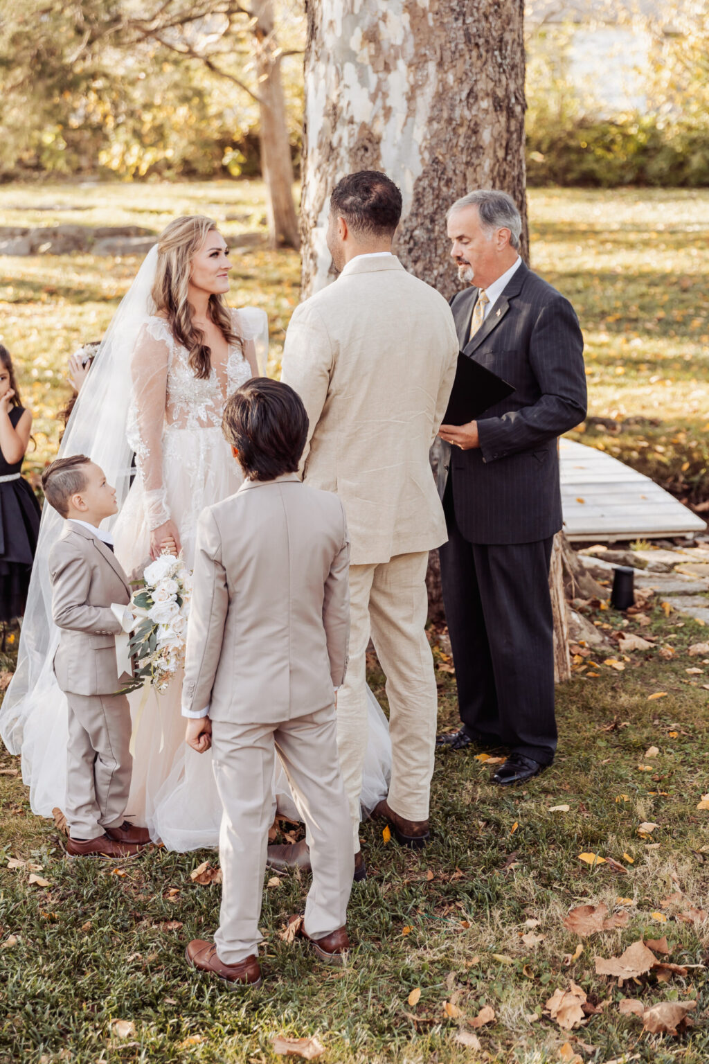 Elopement Wedding Bride and groom stand facing each other at an outdoor elopement ceremony under a large tree. The bride holds a bouquet and wears a white dress with lace details and a long veil. The groom wears a beige suit. Two young boys in beige suits stand nearby, while an officiant in a dark suit stands by the couple. Elopements Inc