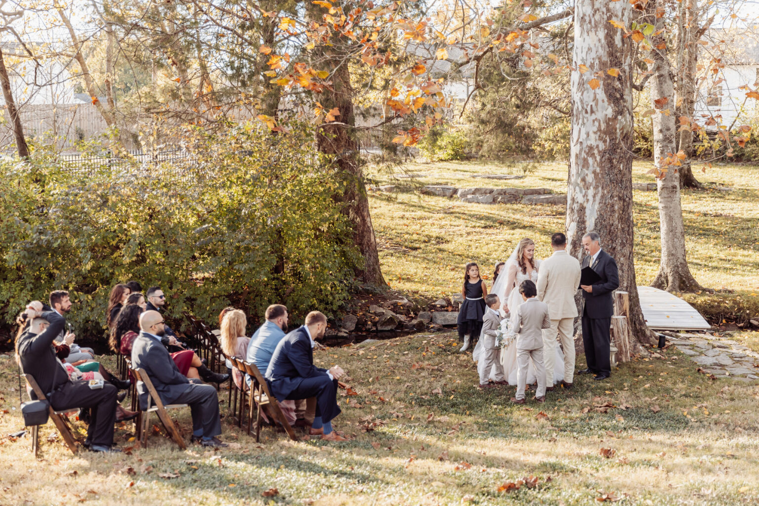 Elopement Wedding A small outdoor wedding ceremony is taking place under a large tree. The bride and groom, who chose to elope, stand facing each other, accompanied by two children. An officiant stands to their right, near a wooden bridge. Guests seated on foldable chairs observe the event amidst autumn foliage and greenery. Elopements Inc