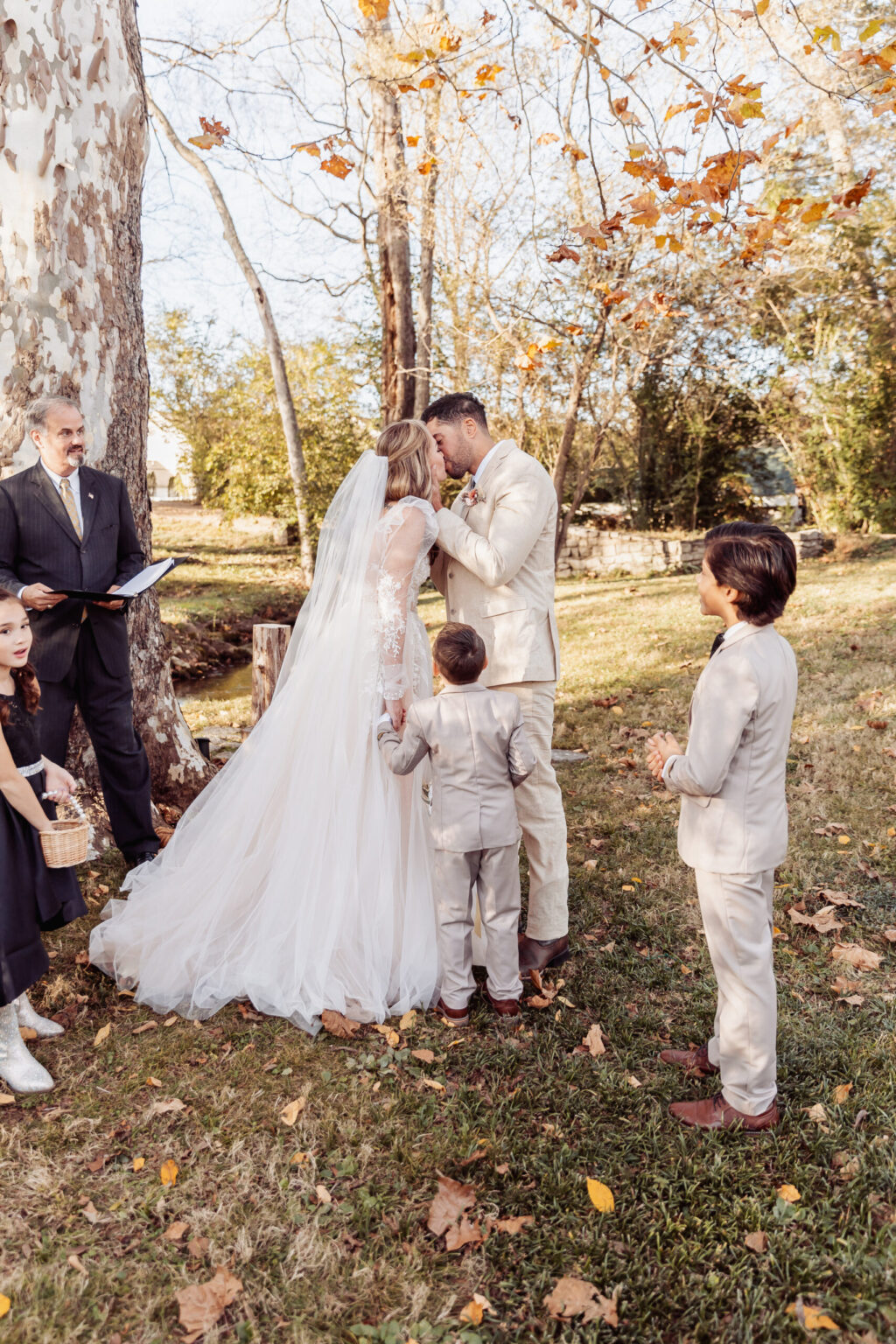 Elopement Wedding A bride and groom share a kiss during their intimate outdoor elopement, surrounded by autumn trees with golden leaves. The bride wears a flowing white gown, and the groom is in a light-colored suit. A young boy, also in a light-colored suit, stands close to them. A minister and other guests are nearby. Elopements Inc