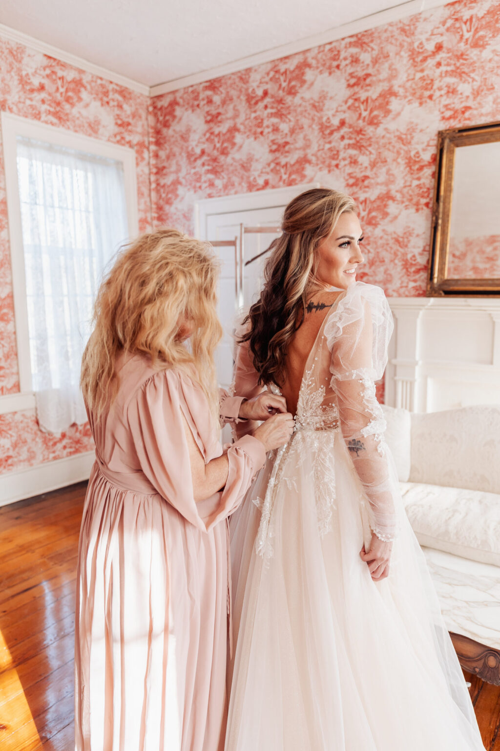 Elopement Wedding A bride in a flowing, lace wedding gown with long sleeves, preparing for her elopement, is assisted by a person with long blonde hair in a light pink dress as they button the back of the dress. They are standing in a room with peach and white floral wallpaper, white trim, and hardwood floors. Elopements Inc