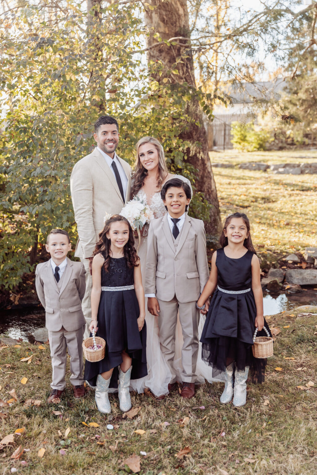 Elopement Wedding A group photo taken outdoors features a couple standing behind four children, all smiling against a backdrop of greenery. Dressed in formal attire, the man and woman radiate joy, possibly celebrating their elopement. The two boys and two girls also wear formal clothes, with the girls holding small baskets. Elopements Inc