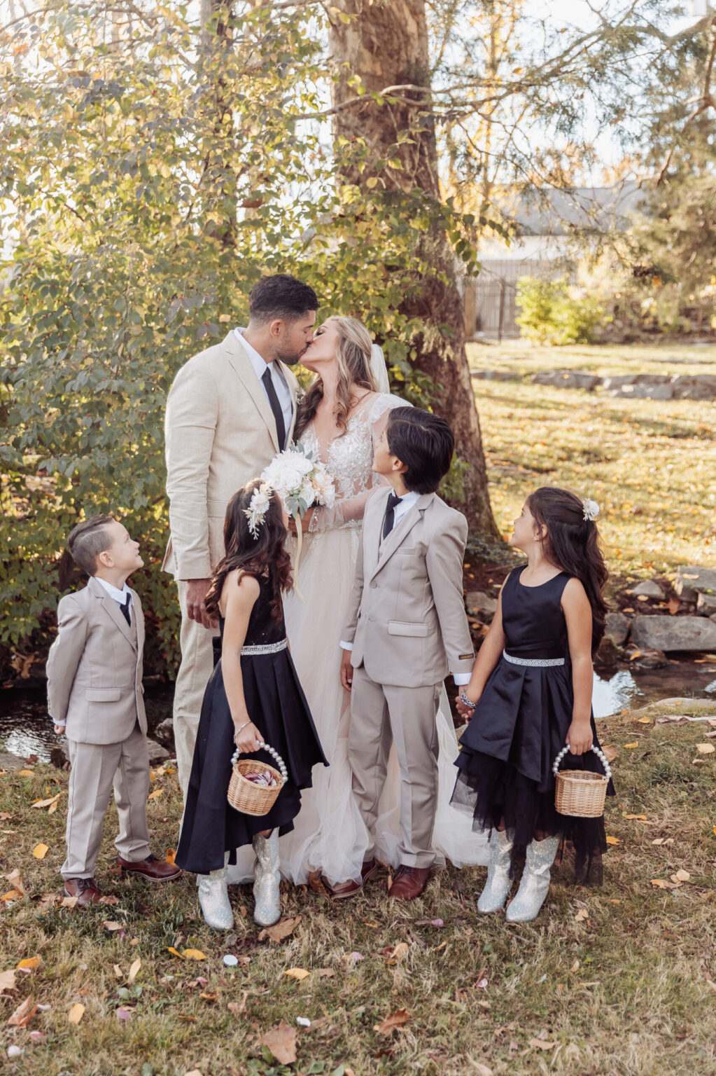 Elopement Wedding Bride and groom sharing a kiss during their elopement, surrounded by five children. The groom is in a light-colored suit, and the bride is in a white wedding dress holding a bouquet. The boys are wearing beige suits, and the girls are in black dresses with white shoes. They are outdoors near a tree and a wooden fence. Elopements Inc