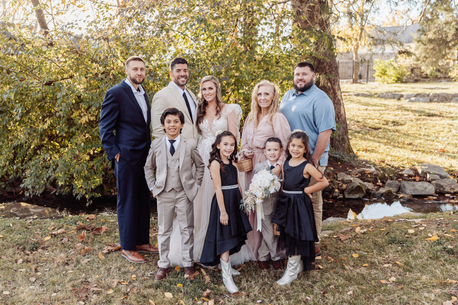 Elopement Wedding A group photo taken outdoors with greenery and trees in the background. Four adults and five children pose together. Two men and a woman on the left are in formal wear, possibly due to an upcoming elopement, while a woman and man on the right are in casual attire. The children are dressed formally, with two girls wearing black dresses. Elopements Inc