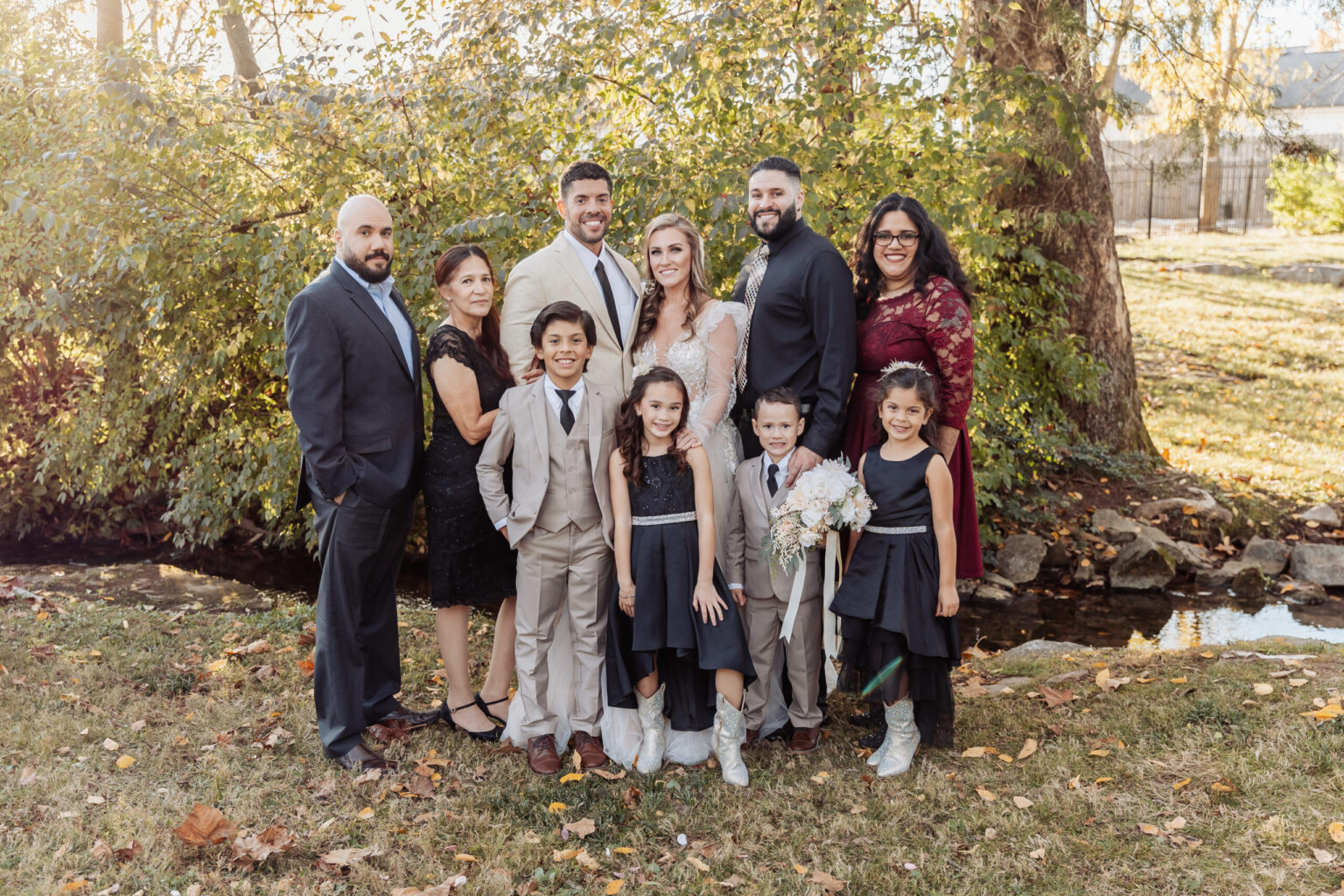 Elopement Wedding A group photo captures ten people standing outdoors. In the center, a bride and groom who recently had an elopement are flanked by family members. There are four children in formal attire, boys in suits and girls in black dresses. The adults are dressed in suits and dresses. Trees and grass create a lush background. Elopements Inc