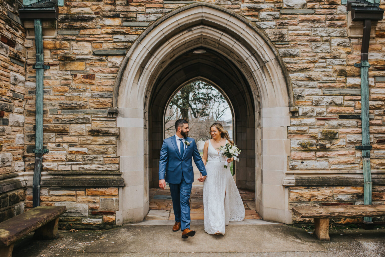 Elopement Wedding A couple walks hand-in-hand under a stone archway. The groom, in a blue suit, gazes at the bride, who wears a white wedding dress and holds a bouquet. They are framed by the textured, light-colored brickwork of the arch, with a worn stone bench to the left and another behind them—an intimate moment of elopement. Elopements Inc