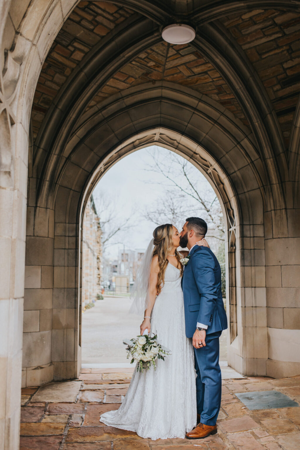 Elopement Wedding A newlywed couple stands under a stone arch, sharing a kiss during their intimate elopement. The bride, in a white lace gown and veil, holds a bouquet of white flowers and greenery. The groom, dressed in a blue suit and brown shoes, gently holds her around the waist. Leafless trees and stone buildings are visible in the background. Elopements Inc