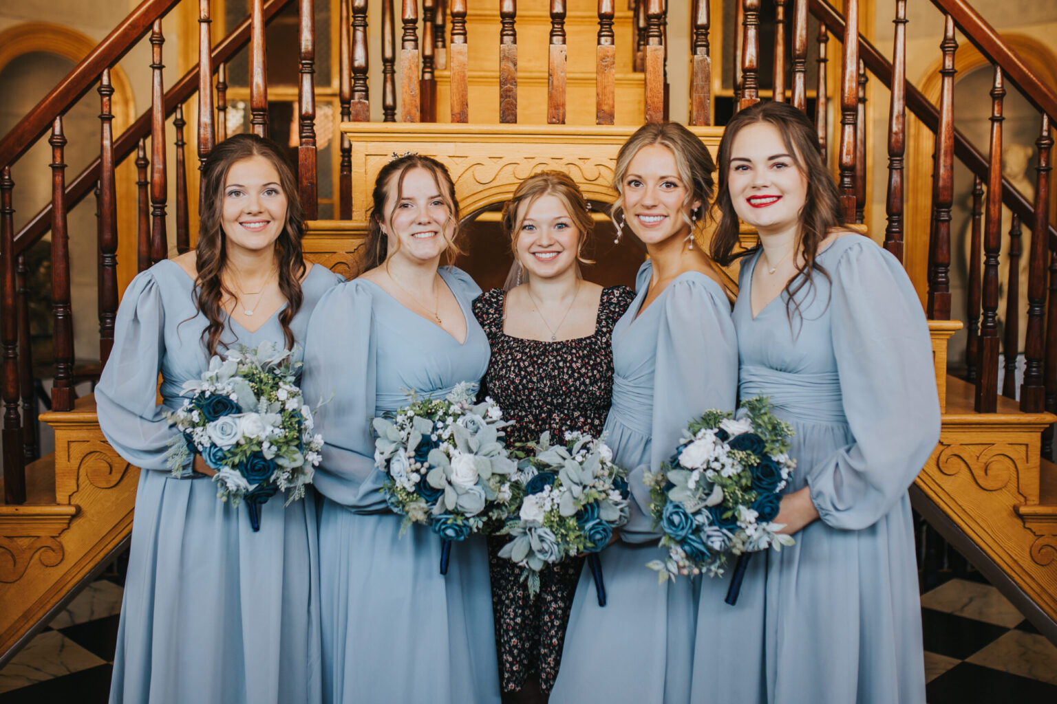 Elopement Wedding Five women stand in front of a wooden staircase. Four of them wear matching light blue dresses and hold bouquets of white and blue flowers. The fifth woman, in the center, wears a black floral-patterned dress and smiles. The background features ornate wooden railings—a perfect setting for an intimate elopement. Elopements Inc