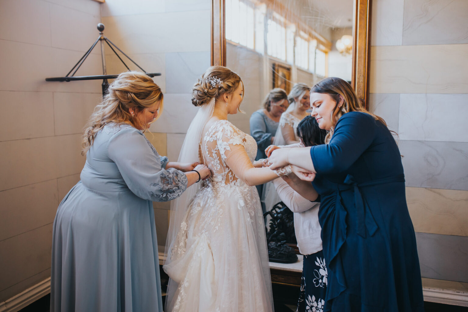 Elopement Wedding A bride in a white lace gown stands in front of a large mirror. Two women assist her with her dress. One, in a light blue dress, adjusts the gown's back, while the other, in a navy dress, works on the sleeve. The serene preparations for their upcoming elopement are reflected softly behind them. Elopements Inc