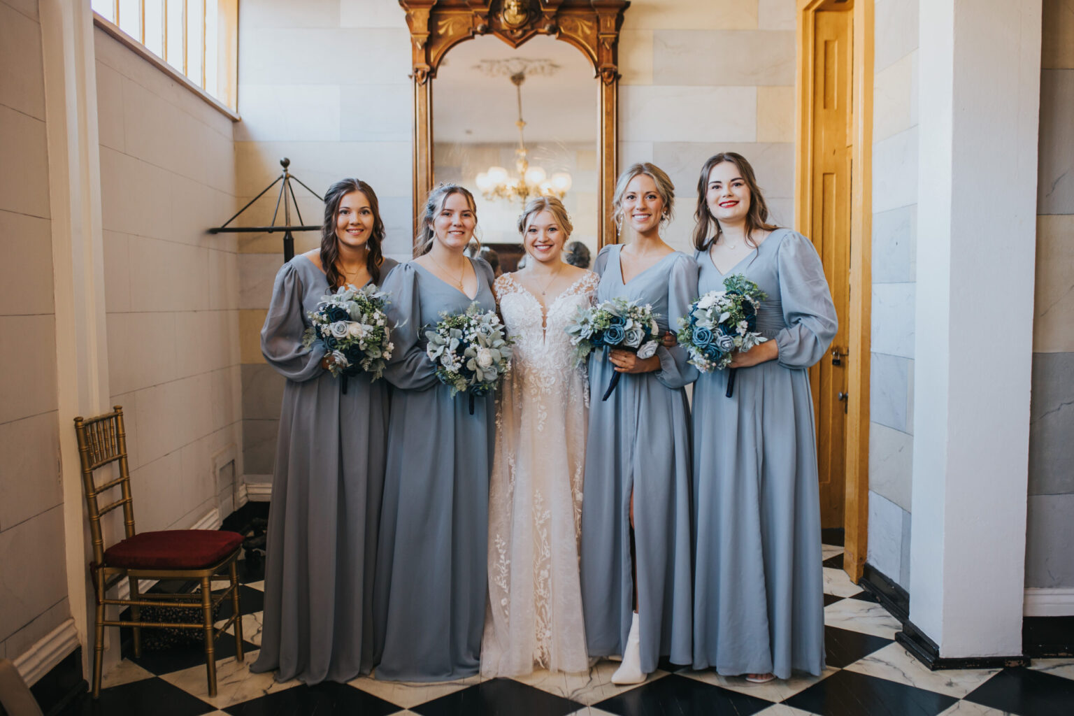 Elopement Wedding A bride in a white wedding dress stands with four bridesmaids wearing light blue dresses, all holding bouquets of blue and white flowers. They are posing indoors in front of a wooden-framed mirror with a visible chandelier, after deciding to elope. A wooden chair with a red seat is on the left side of the image. Elopements Inc