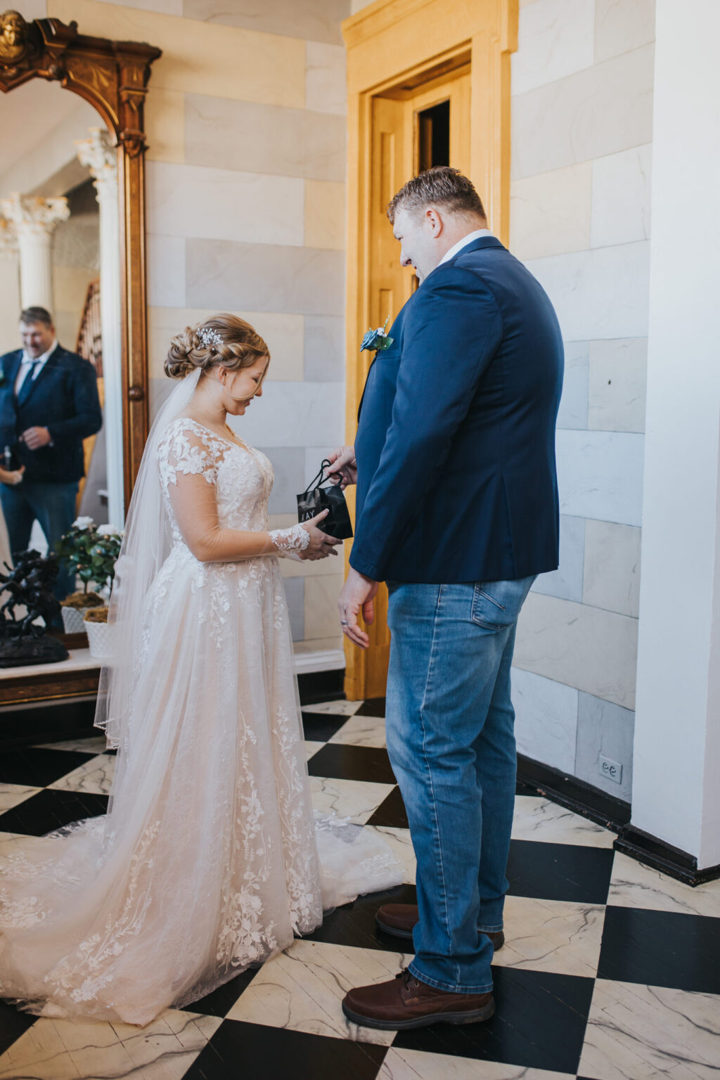 Elopement Wedding A bride in a white lace gown and a groom in a blue blazer and jeans are standing indoors on a checkered black-and-white floor. The groom is placing a ring on the bride's finger. A man with a beard, wearing a suit, stands in the background, observing the intimate elopement moment. Elopements Inc