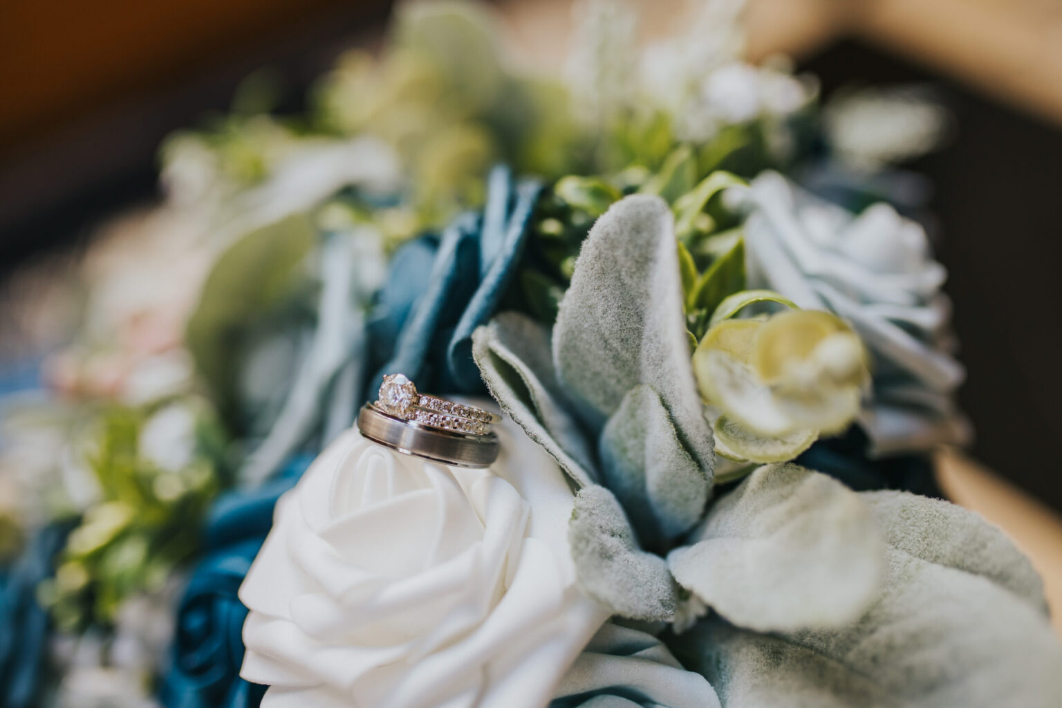 Elopement Wedding A close-up shot of two rings on a bouquet of artificial flowers, perfect for elopements. The rings, one with a diamond and one plain band, rest on white and grey fabric flowers. The background is softly blurred with hints of green foliage. Elopements Inc