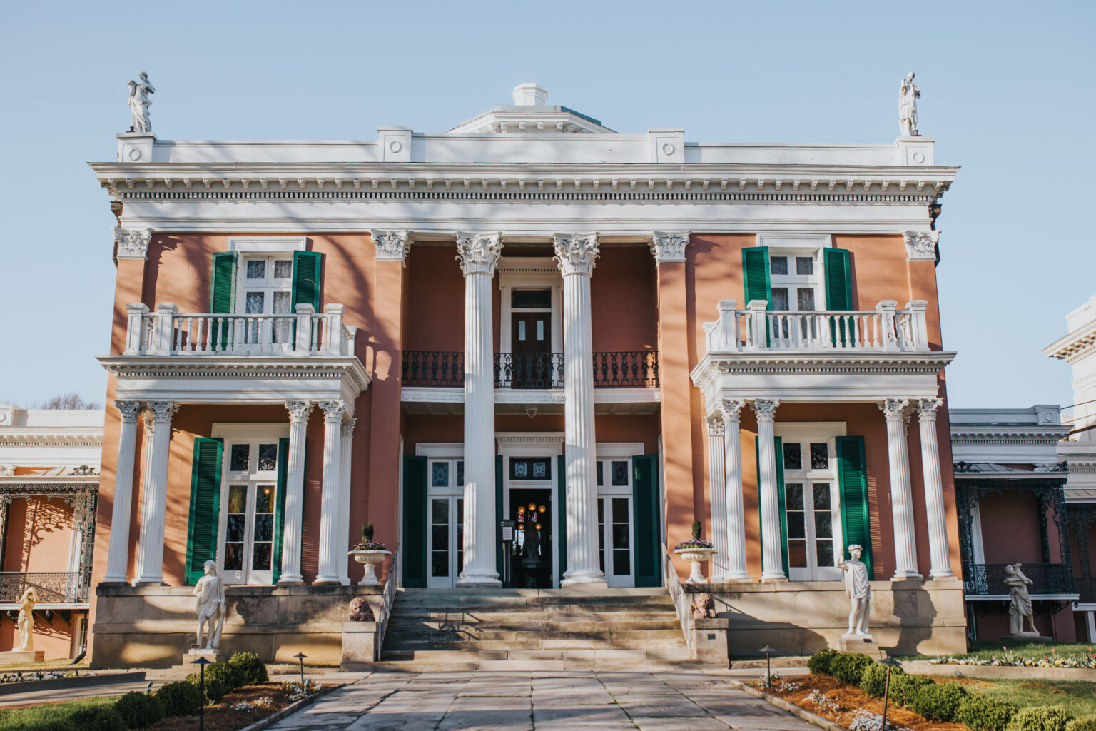 Elopement Wedding A stately building with a pink façade and white trim features grand columns at its entrance. Two upper balconies with green shutters and statues adorn the front. Manicured landscaping and a stone pathway lead to the main doors. It's an idyllic setting for an elopement, basked in clear sunlight. Elopements Inc