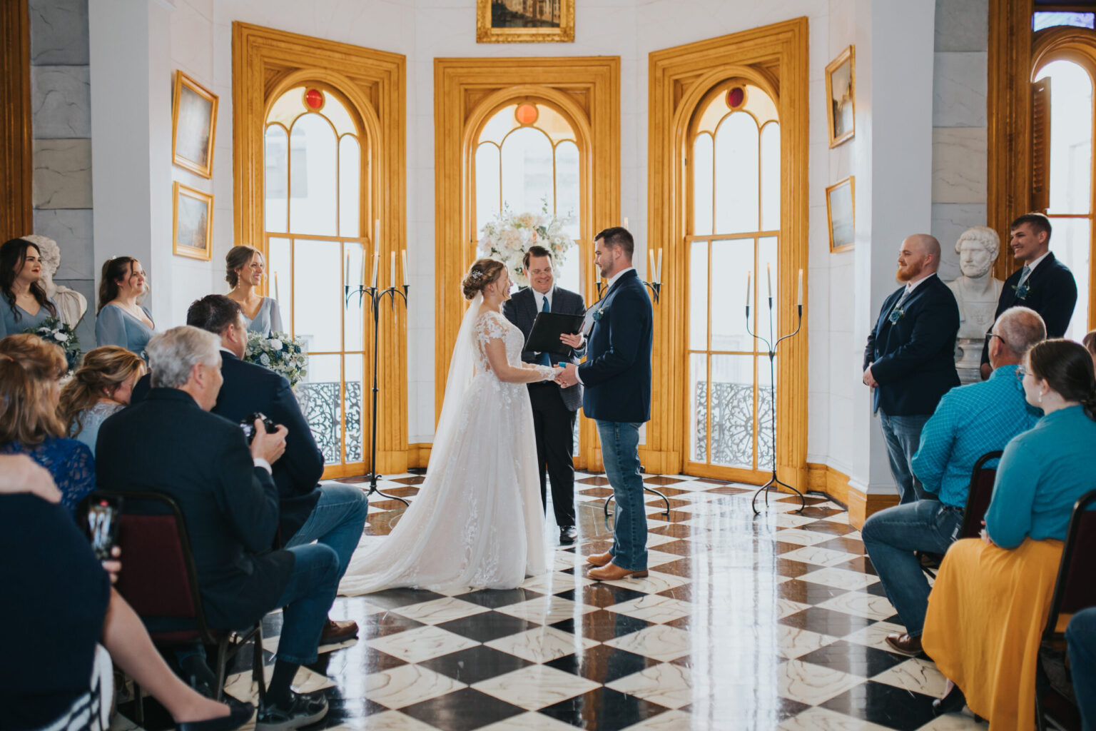 Elopement Wedding A couple stands before each other, holding hands, during their wedding ceremony in an ornate room with arched, golden-framed windows. The bride wears a white gown with a long train, and the groom is in a dark suit. Unlike traditional elopements, guests are seated, watching the ceremony and capturing the moment with their phones. Elopements Inc