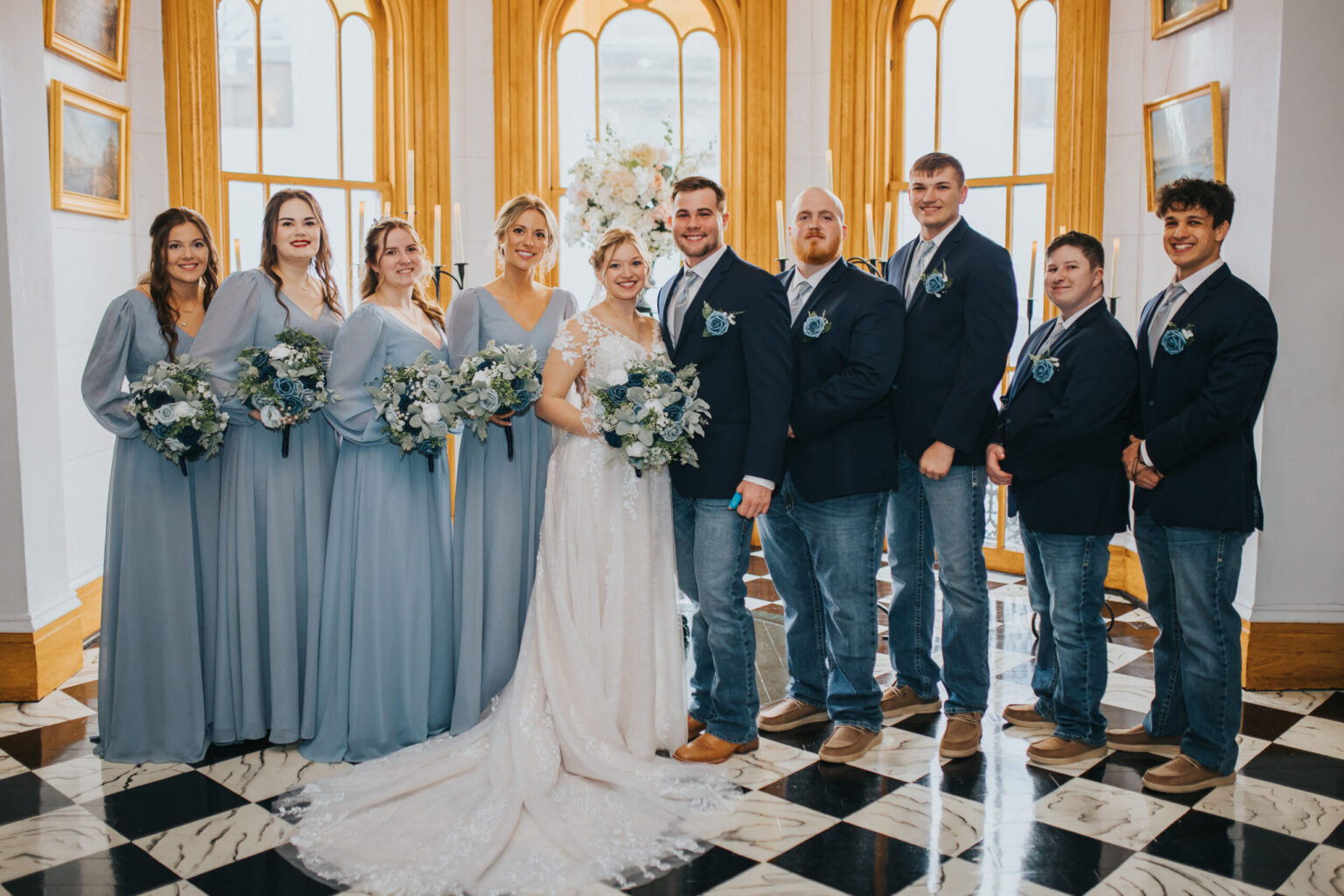 Elopement Wedding A wedding party poses in front of large, arched windows. The bride, in a white gown, stands at the center arm-in-arm with the groom, who wears a dark jacket and jeans. Bridesmaids in light blue dresses hold bouquets, while groomsmen in dark jackets and jeans stand on either side—a beautiful scene for an elopement. Elopements Inc