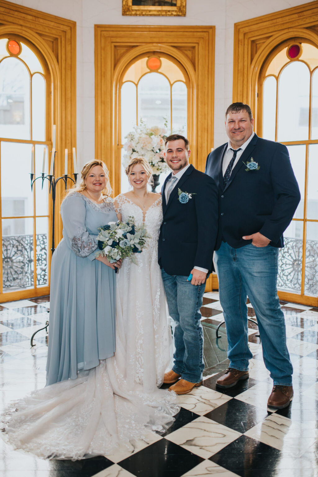 Elopement Wedding Four people are posing in a brightly lit room with large windows and a marble floor. The bride, in a lace gown and holding a bouquet, stands beside the groom in a navy suit and jeans. Two other people are on either side, both in light blue dresses and a navy suit with jeans, capturing an intimate elopement moment. Elopements Inc