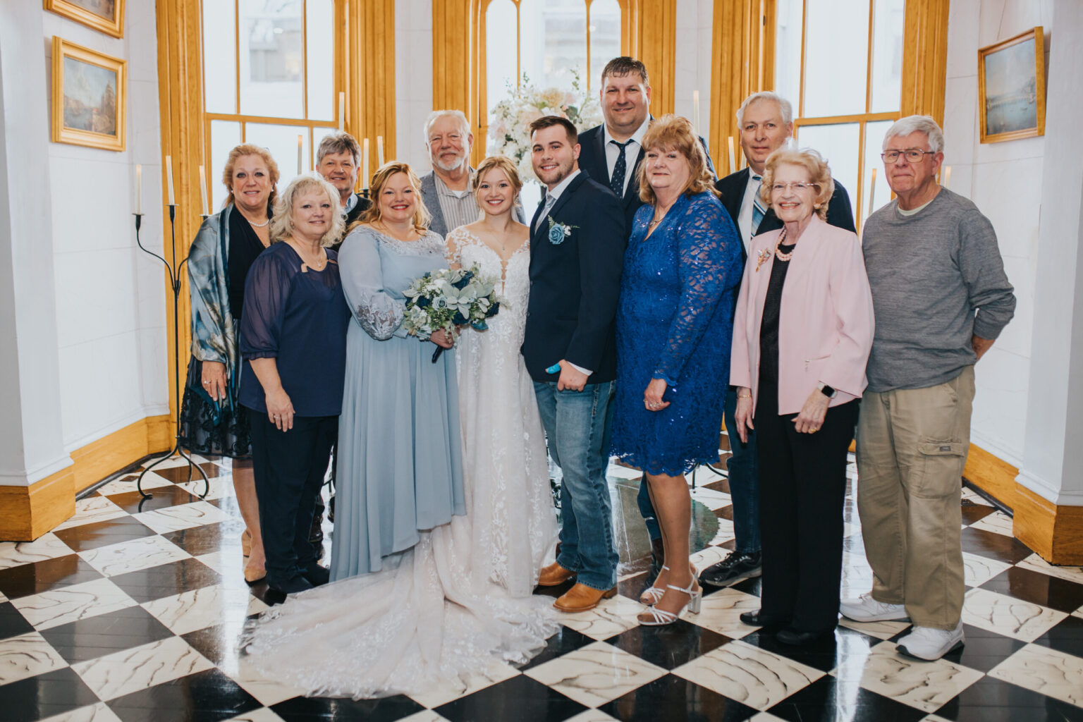 Elopement Wedding A wedding group photo features a bride in a lace gown holding a bouquet, and a groom in a navy suit standing at the center. They are surrounded by family members and friends, all dressed in semi-formal attire. The setting has large windows and checkered black-and-white flooring, perfect for those who chose not to elope. Elopements Inc