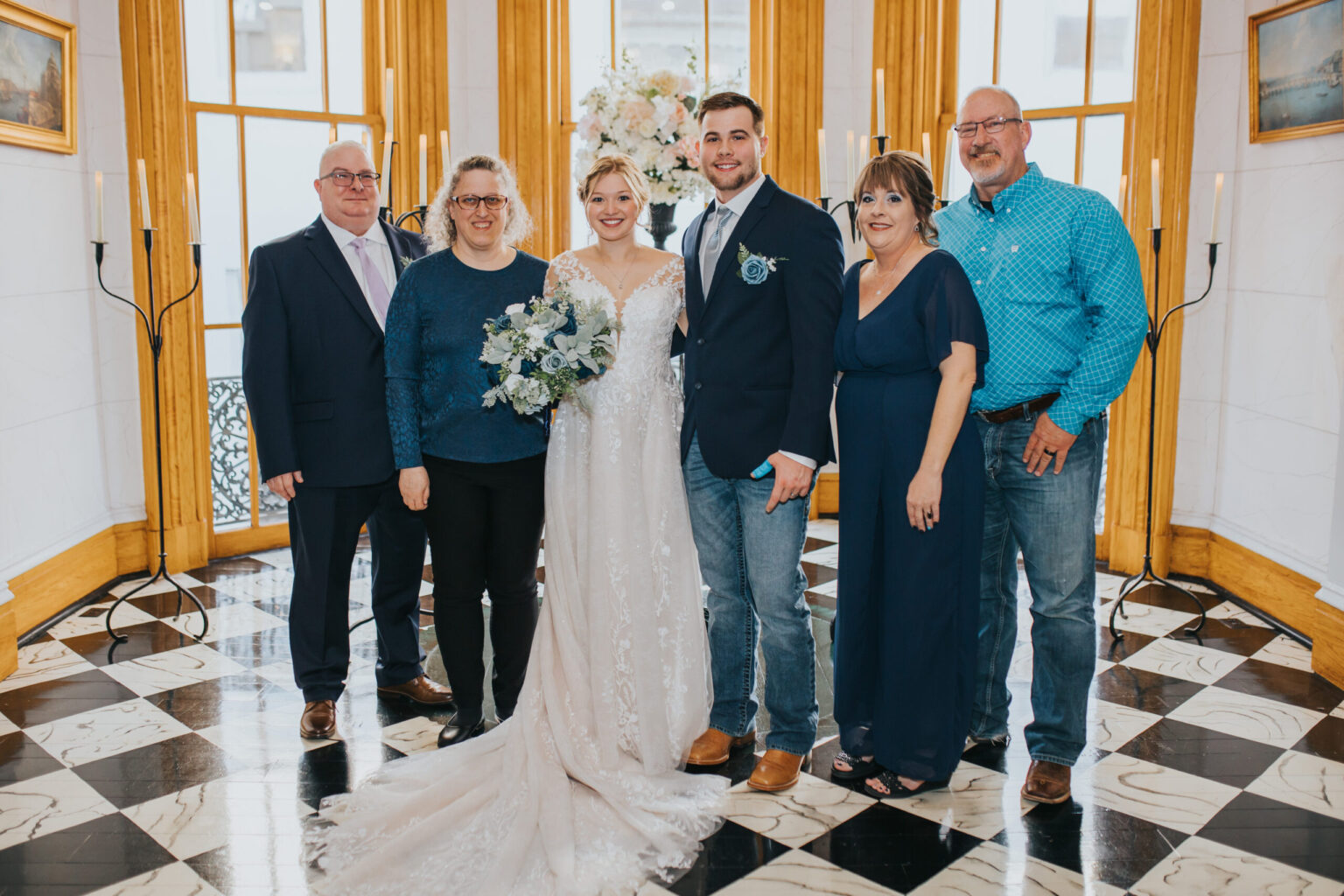 Elopement Wedding A bride in a lace gown and a groom in a dark suit, who decided to elope, stand in the center of a group flanked by three other adults. The group is in an elegant room with black and white checkered floors, large windows, golden curtains, and lit candles. They are all dressed formally, smiling at the camera. Elopements Inc