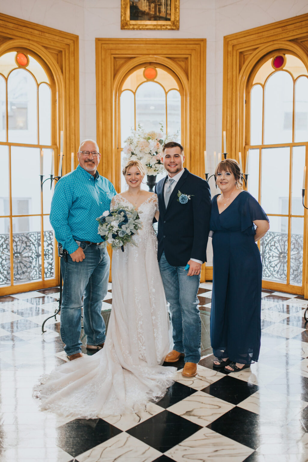 Elopement Wedding A bride and groom who chose to elope stand in the center, flanked by an older man and woman. The bride wears a white gown and holds a bouquet, while the groom is dressed in a dark suit with a tie. The older man wears a blue shirt and jeans, and the older woman wears a dark blue dress. They are in an elegant room with large arched windows and a Elopements Inc