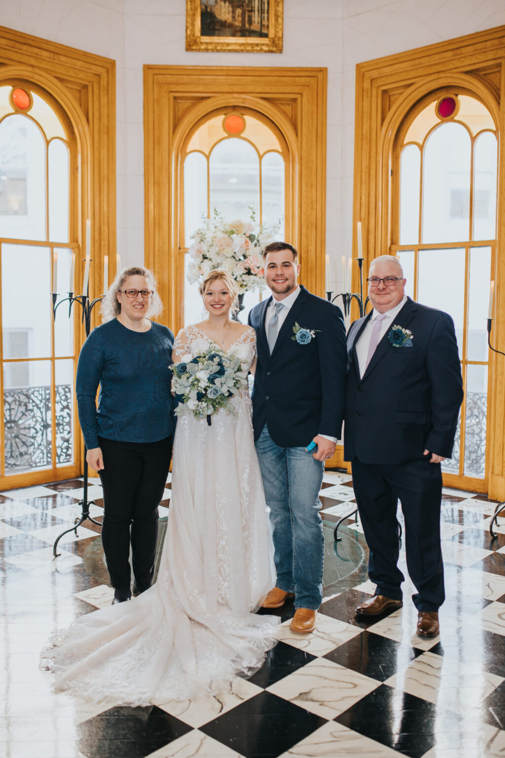 Elopement Wedding A bride in a white gown and floral headdress stands beside a groom in jeans and a suit jacket, flanked by two other individuals, one in a blue sweater and the other in a navy suit. They are celebrating their elopement in an ornate room with large arched windows, black-and-white checkered flooring, and candelabras. Elopements Inc