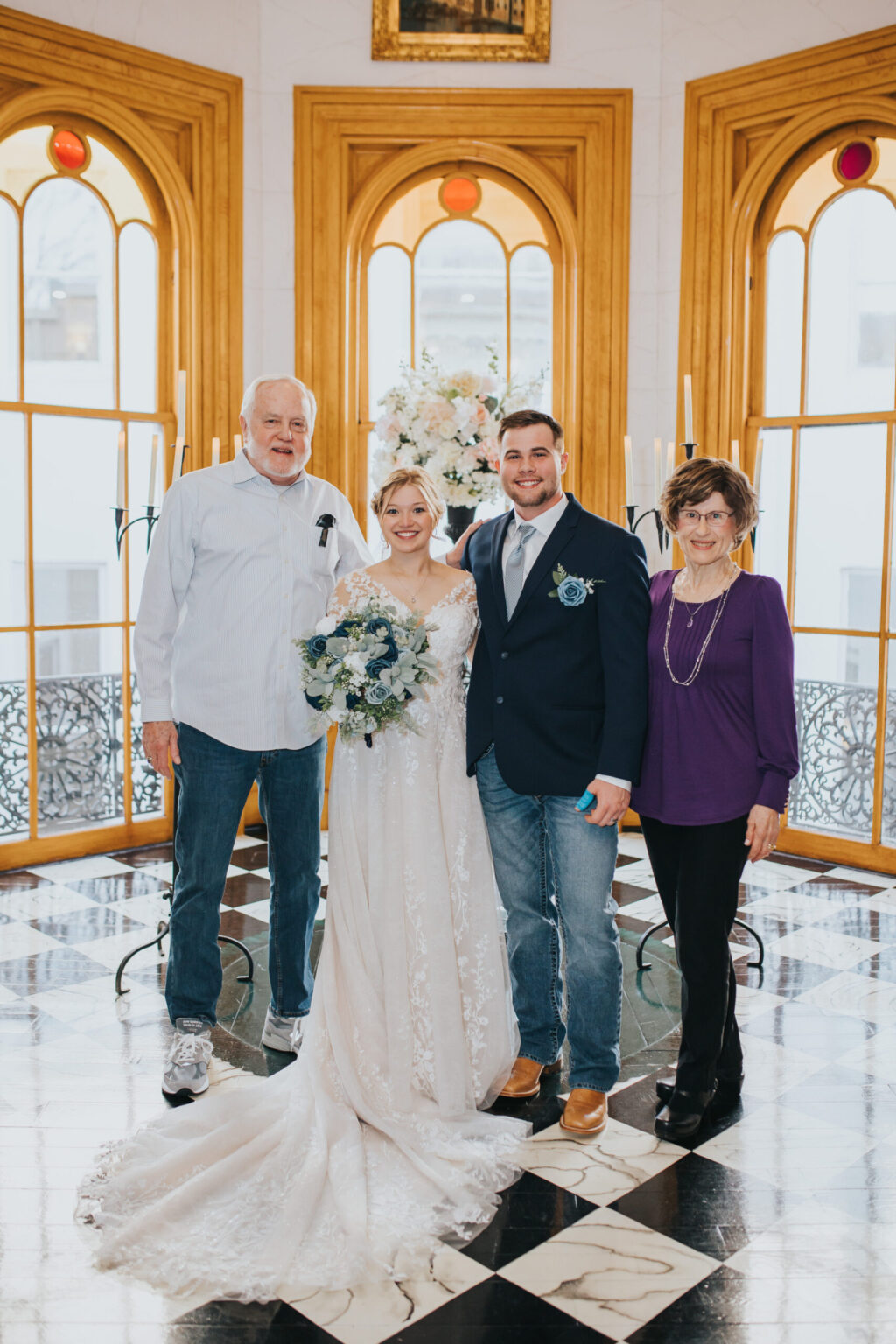 Elopement Wedding A wedding photo shows four people standing together inside a beautifully decorated room with large arched windows and checkered floor tiles. The bride, having just eloped, is in a white dress holding a bouquet and standing next to the groom in a blue blazer and jeans. An older couple stands on either side of them. Elopements Inc