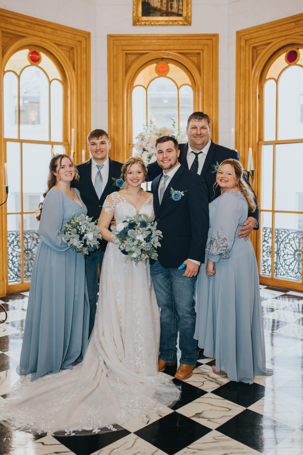 Elopement Wedding A wedding portrait in a grand setting with large arched windows. Center, the bride is in a white gown holding a bouquet and the groom in a dark suit with jeans. Flanked by two bridesmaids in blue dresses and two groomsmen in suits with blue ties. Everyone is smiling warmly—an elegant touch to an intimate elopement. Elopements Inc