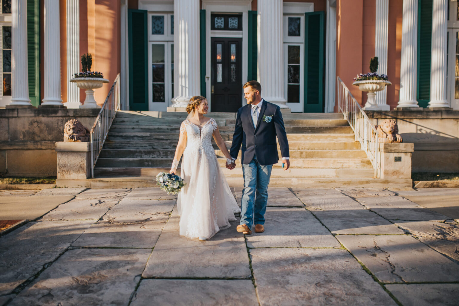 Elopement Wedding A couple in wedding attire holds hands while walking in front of a grand building with tall white columns and green shutters. The bride wears a white gown and carries a bouquet, and the groom is dressed in a dark blazer, light shirt, and jeans. Stone steps and potted plants frame the charming elopement entrance. Elopements Inc