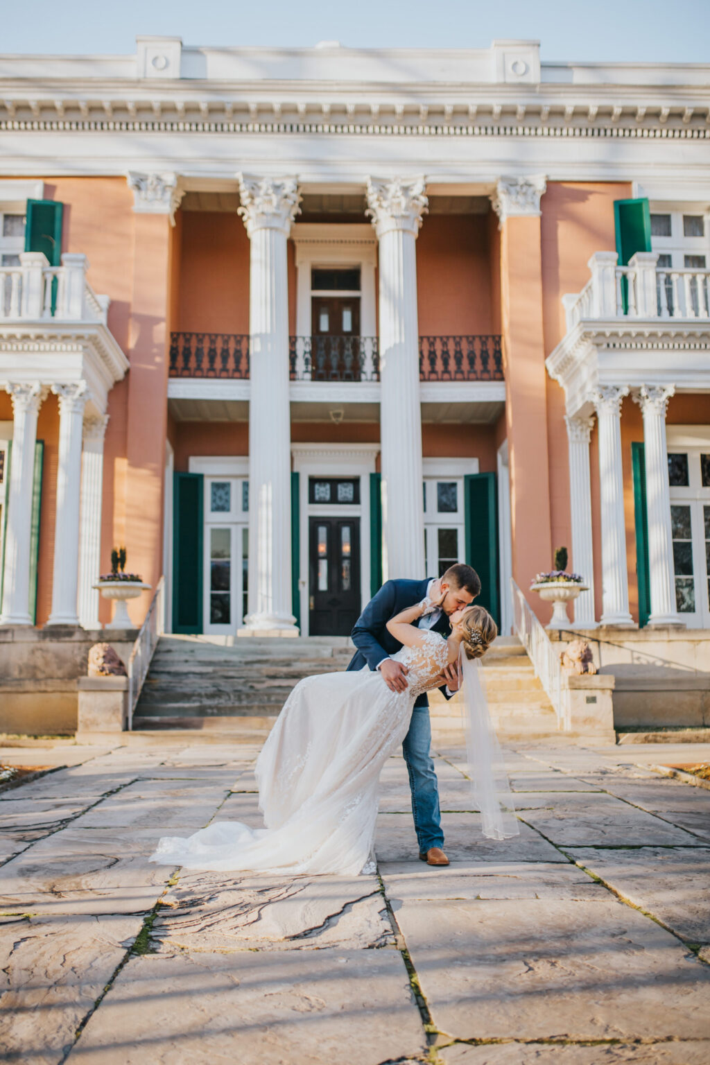 Elopement Wedding A bride and groom share a romantic kiss in front of a grand, historic building with white columns and pale pink walls. The bride, in a flowing white gown and veil, and the groom, in a dark suit, are standing on an expansive stone courtyard, bathed in warm sunlight. Their intimate elopement feels magical. Elopements Inc