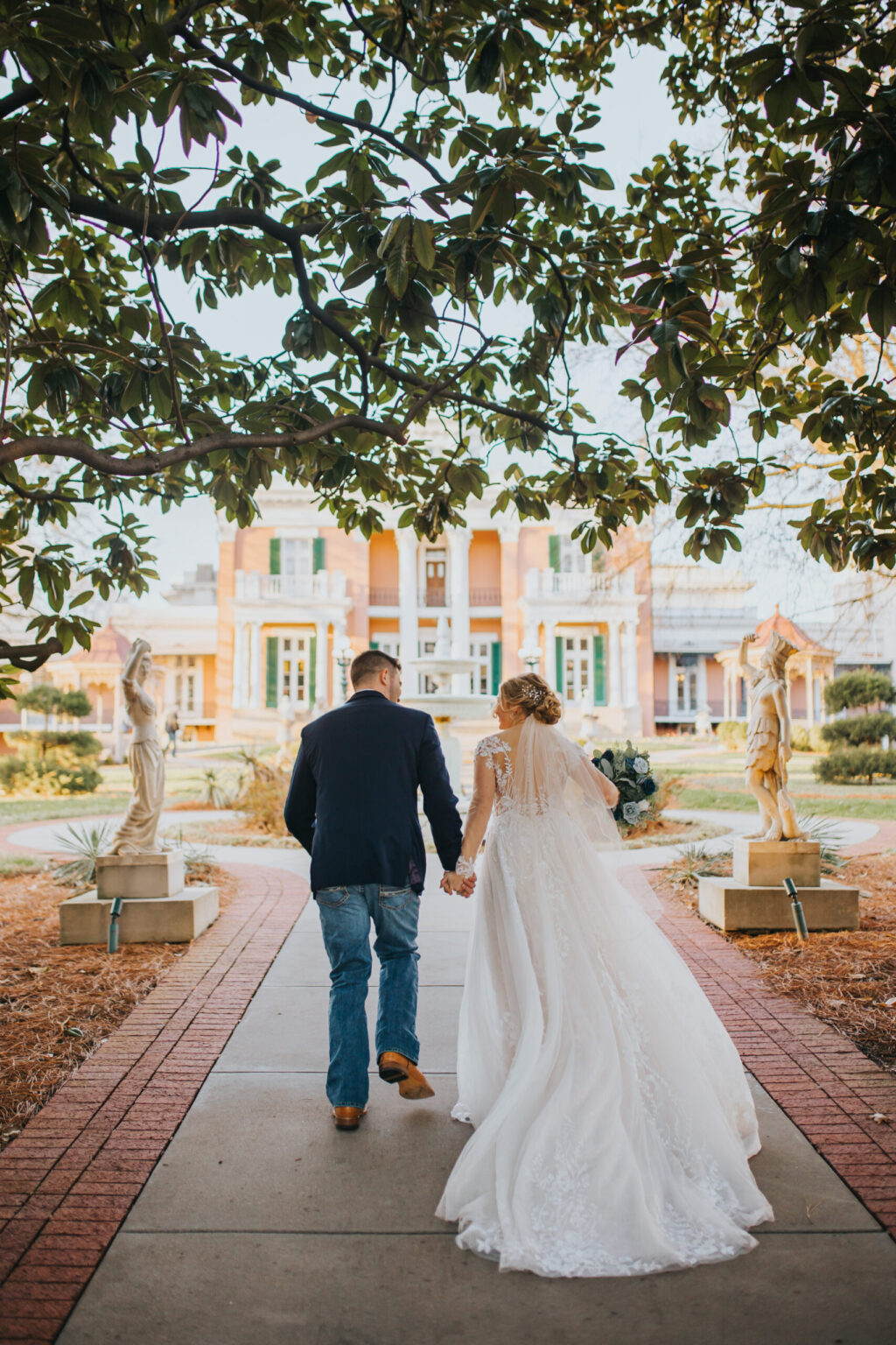 Elopement Wedding A bride and groom walk hand-in-hand down a brick-lined path toward a large, stately historic building. The bride wears a flowing white gown, and the groom is in a dark jacket and jeans. Statues and greenery frame the path, while a large, overhanging tree provides a natural canopy above them on their elopement journey. Elopements Inc