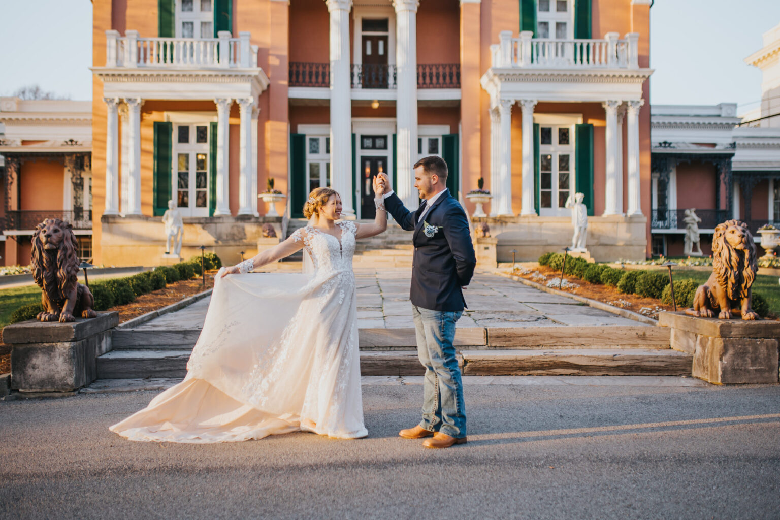 Elopement Wedding A couple, eloping in secret, dances in front of a grand, salmon-colored building with white columns. The woman wears a flowing, white wedding dress, and the man is dressed in a dark blazer, jeans, and boots. Stone lion statues flank the steps leading to the building’s entrance, adorned with intricate details. Elopements Inc